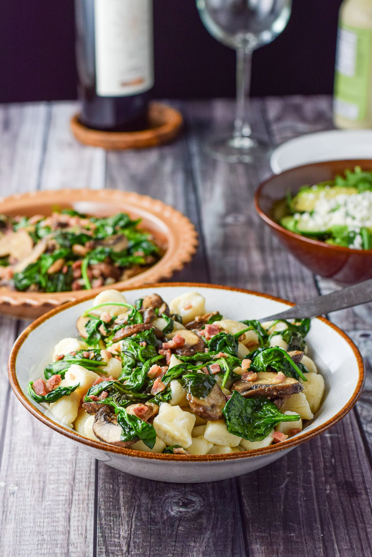 A white bowl with the gnocchi dish in it with a plate with the vegetable in it along with a salad and wine