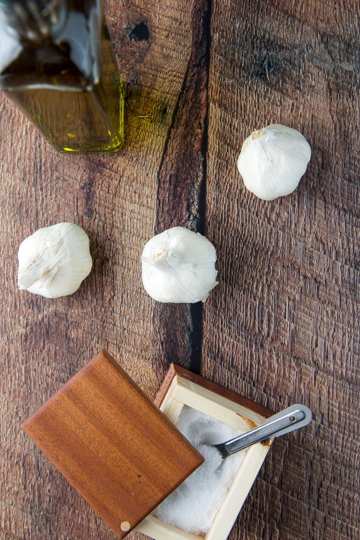 Overhead view of three heads of garlic, salt in a container and olive oil