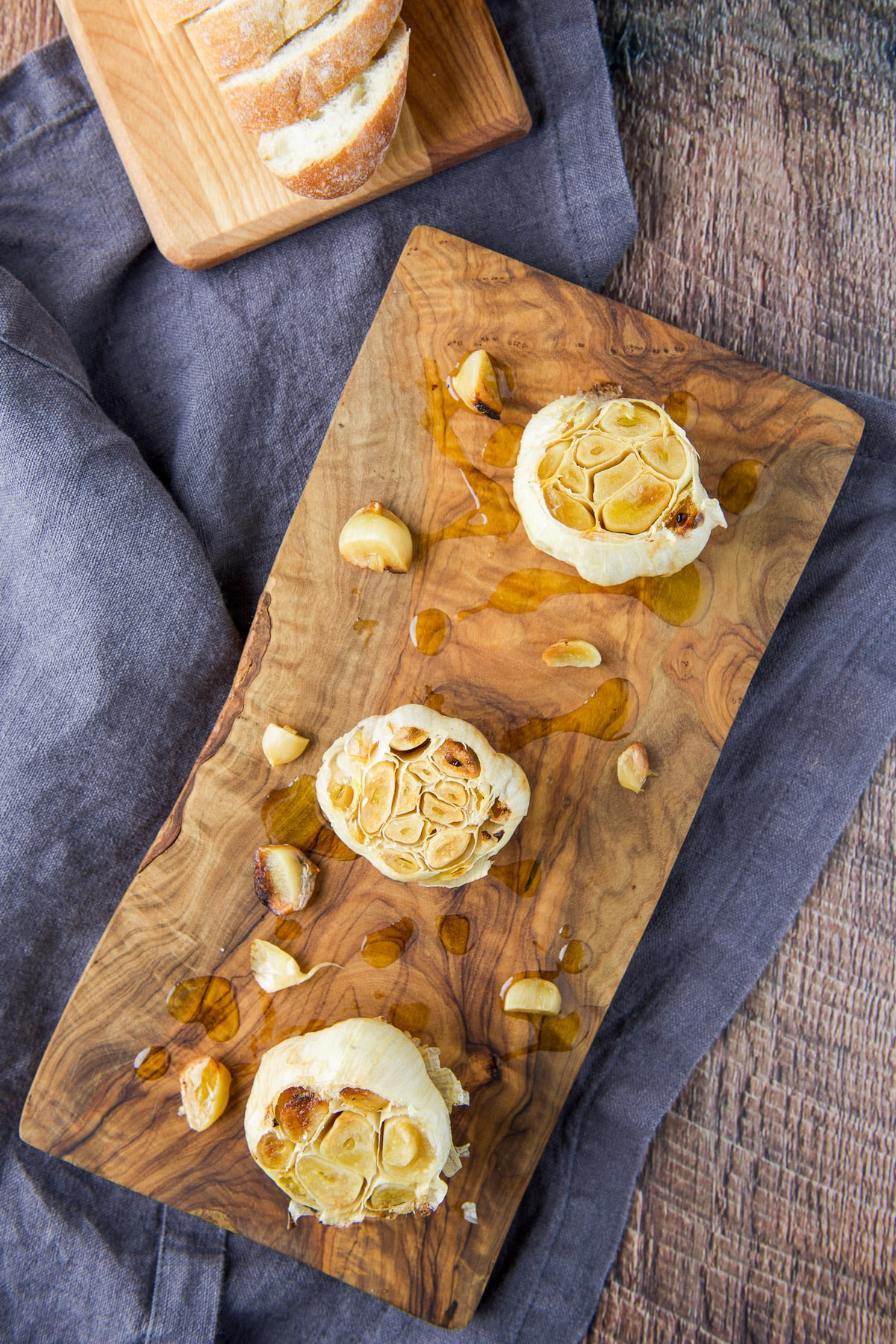 Overhead view of a wood boards one with three roasted garlic heads and one with bread