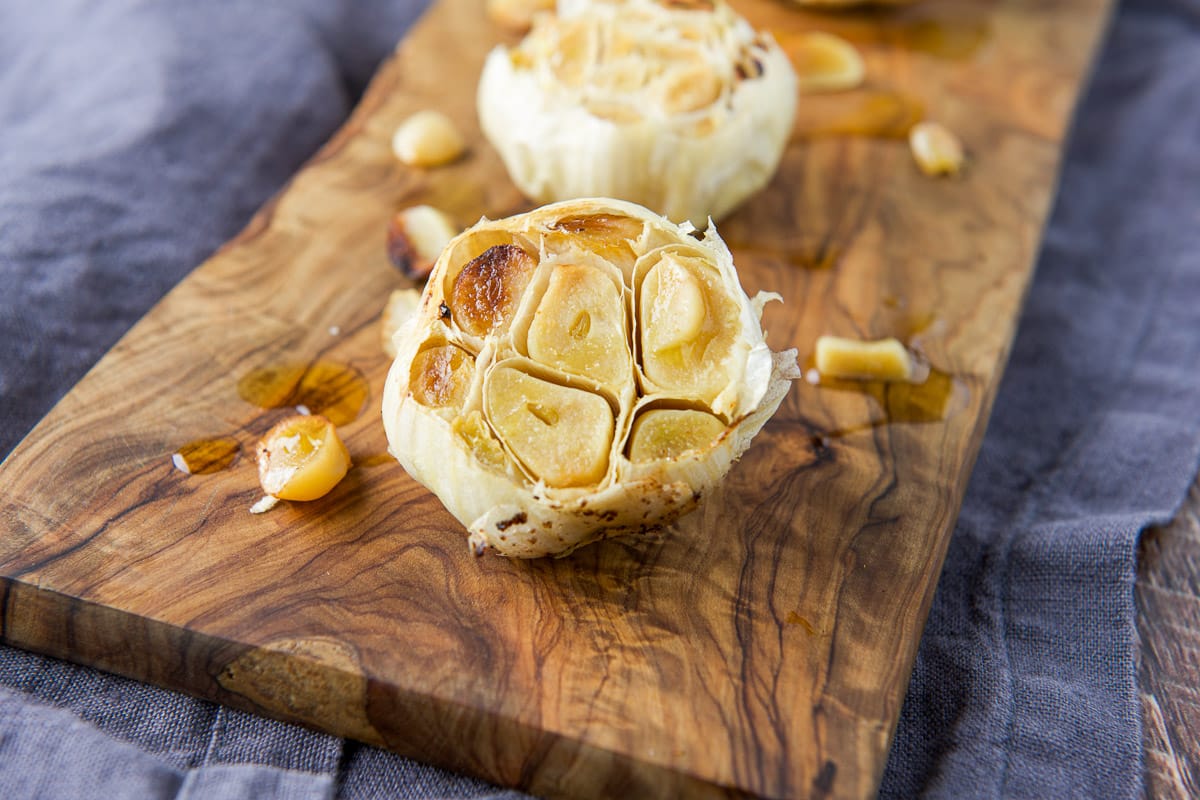 horizontal photo of cooked garlic heads on a board with oil and single cloves