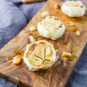 square photo of a wood board with roasted garlic head on it along with single cloves