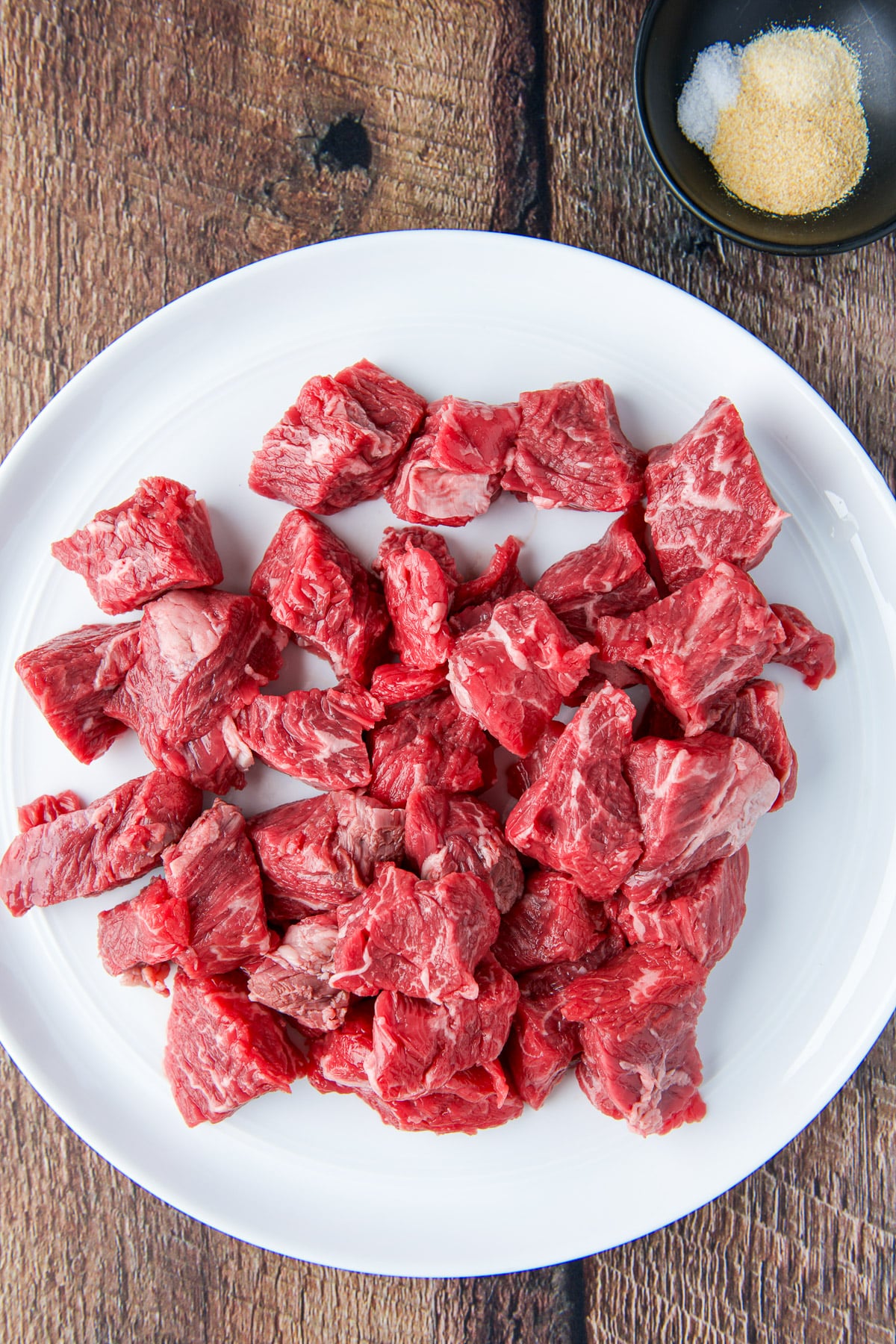 Overhead view of a white plate with raw steak tips on it along with a black bowl with garlic and onion powder on it along with salt