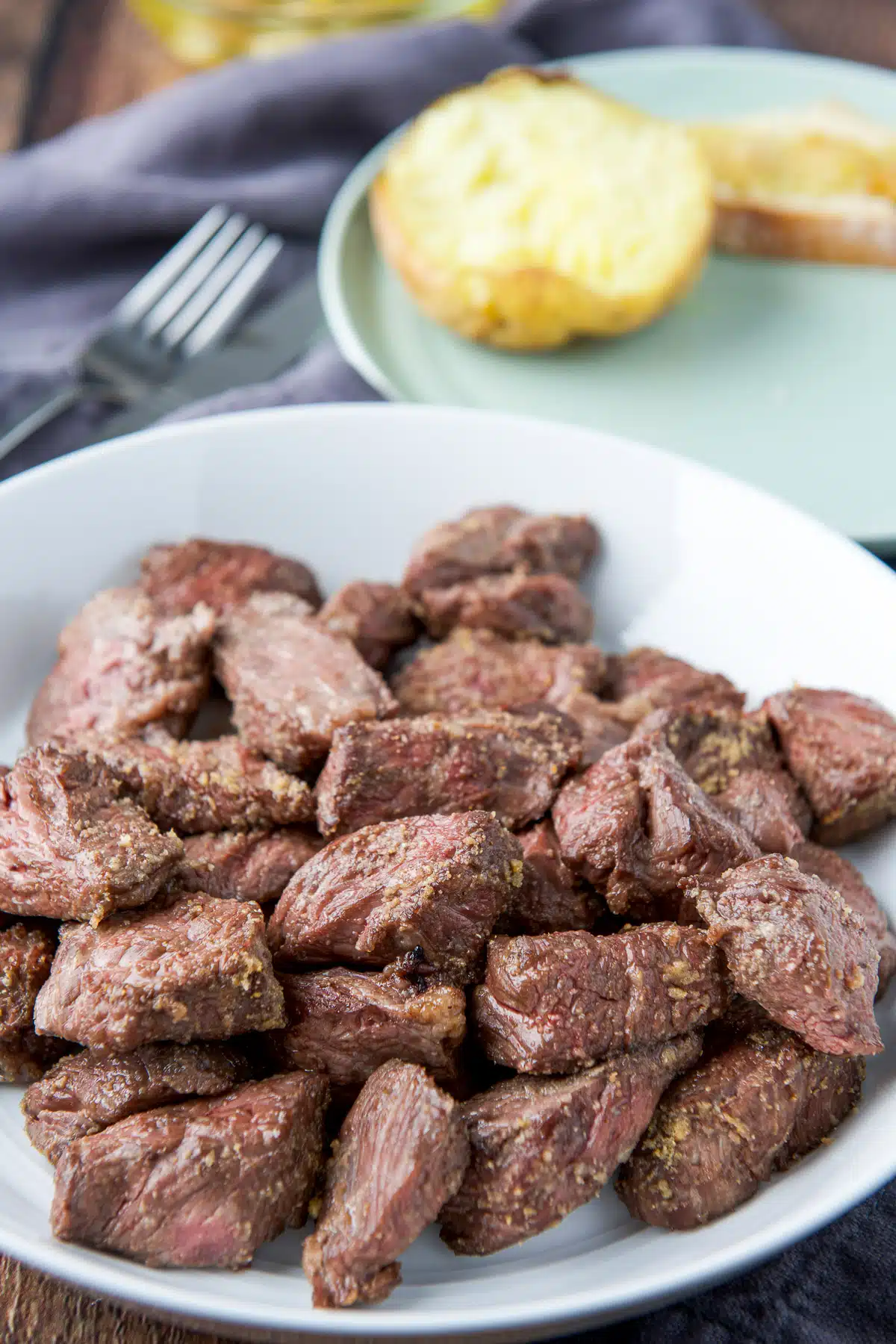 A white deep dish with steak tips in it along with a plate with a potato and bread