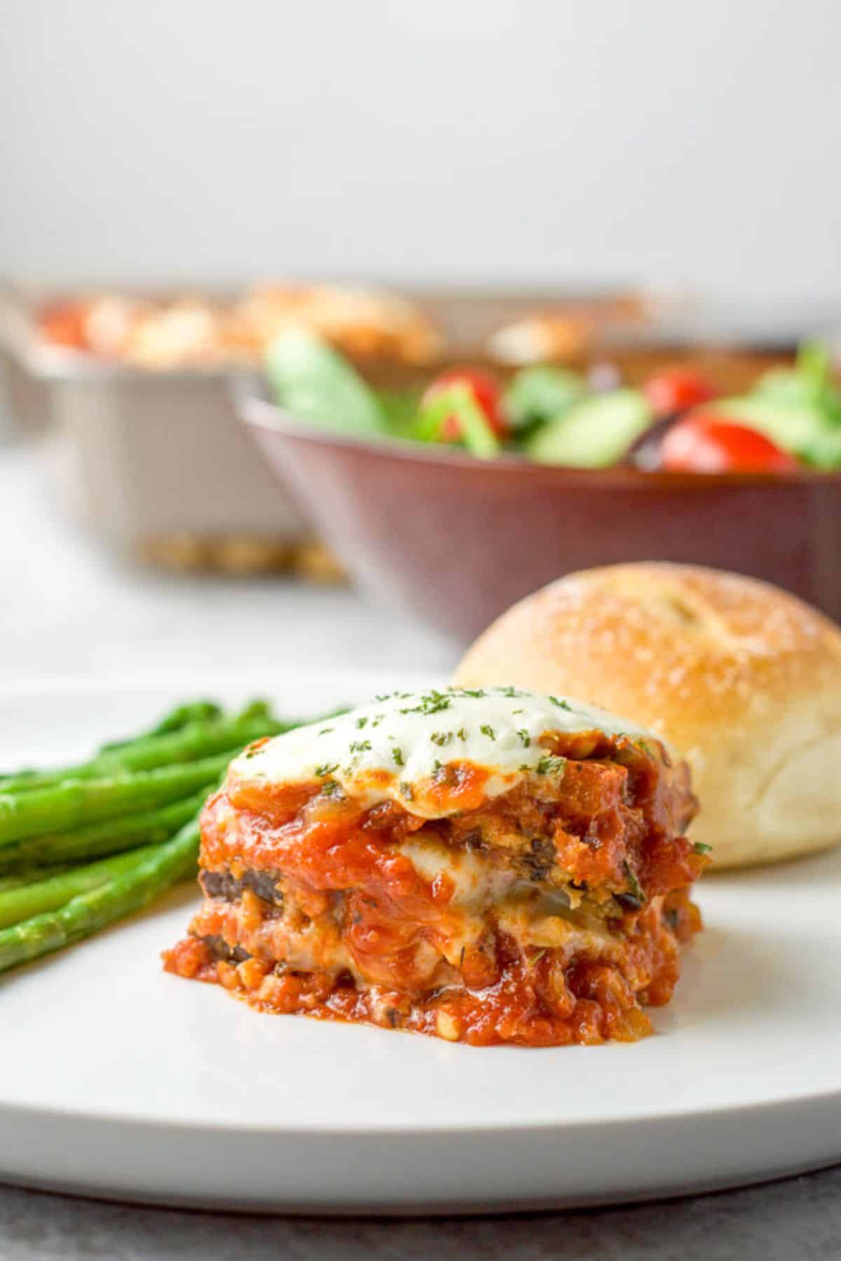 A white plate with the eggplant parm, roll, and asparagus along with a salad and the pan of eggplant in the back