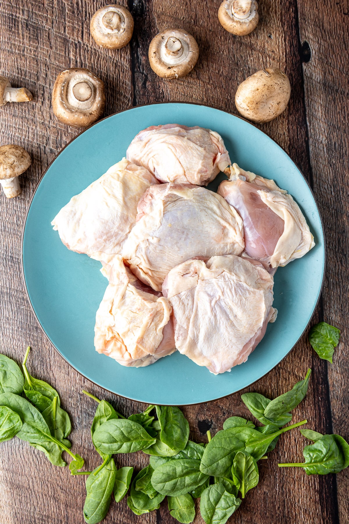 Overhead view of mushrooms, chicken thighs on a blue plate, and spinach on a table