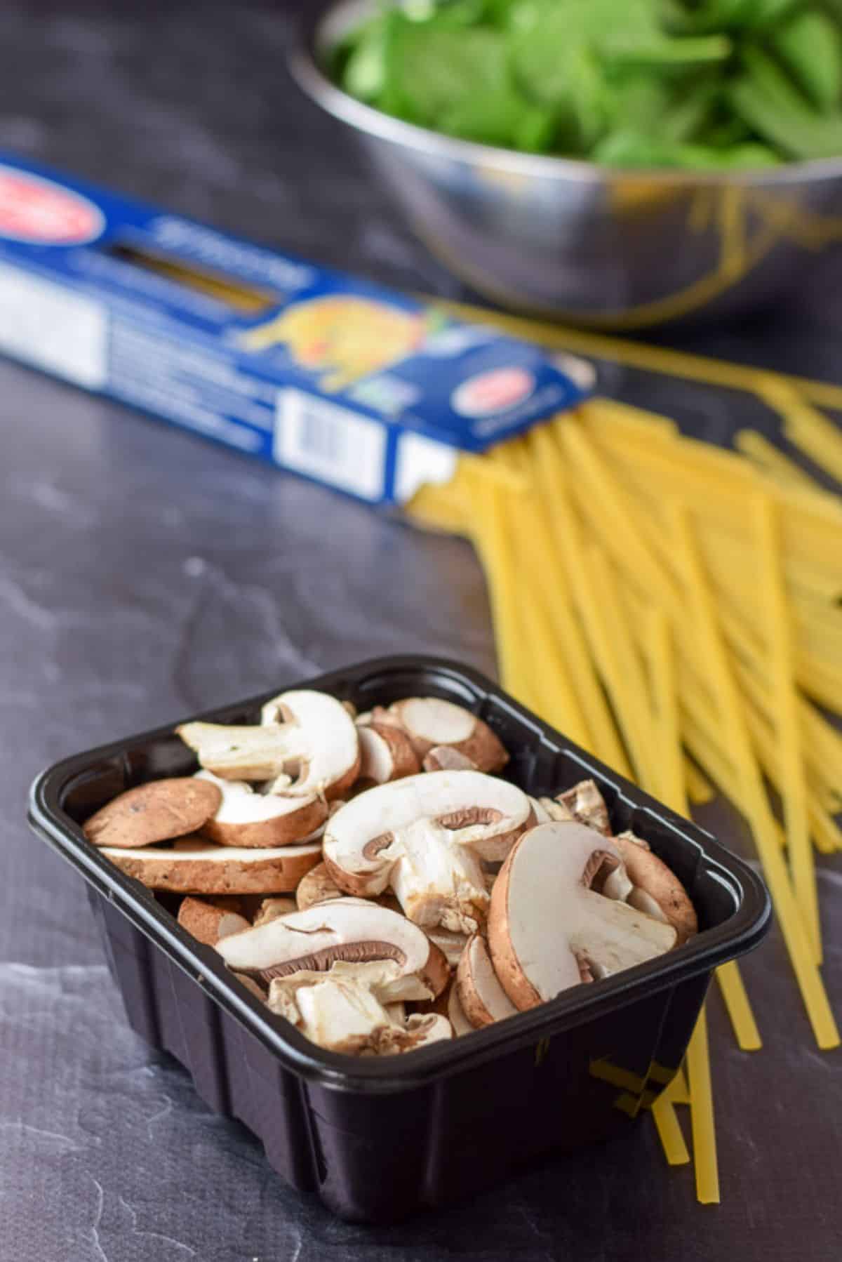 A container of sliced mushrooms, fettuccine strewn on the table and spinach in a bowl