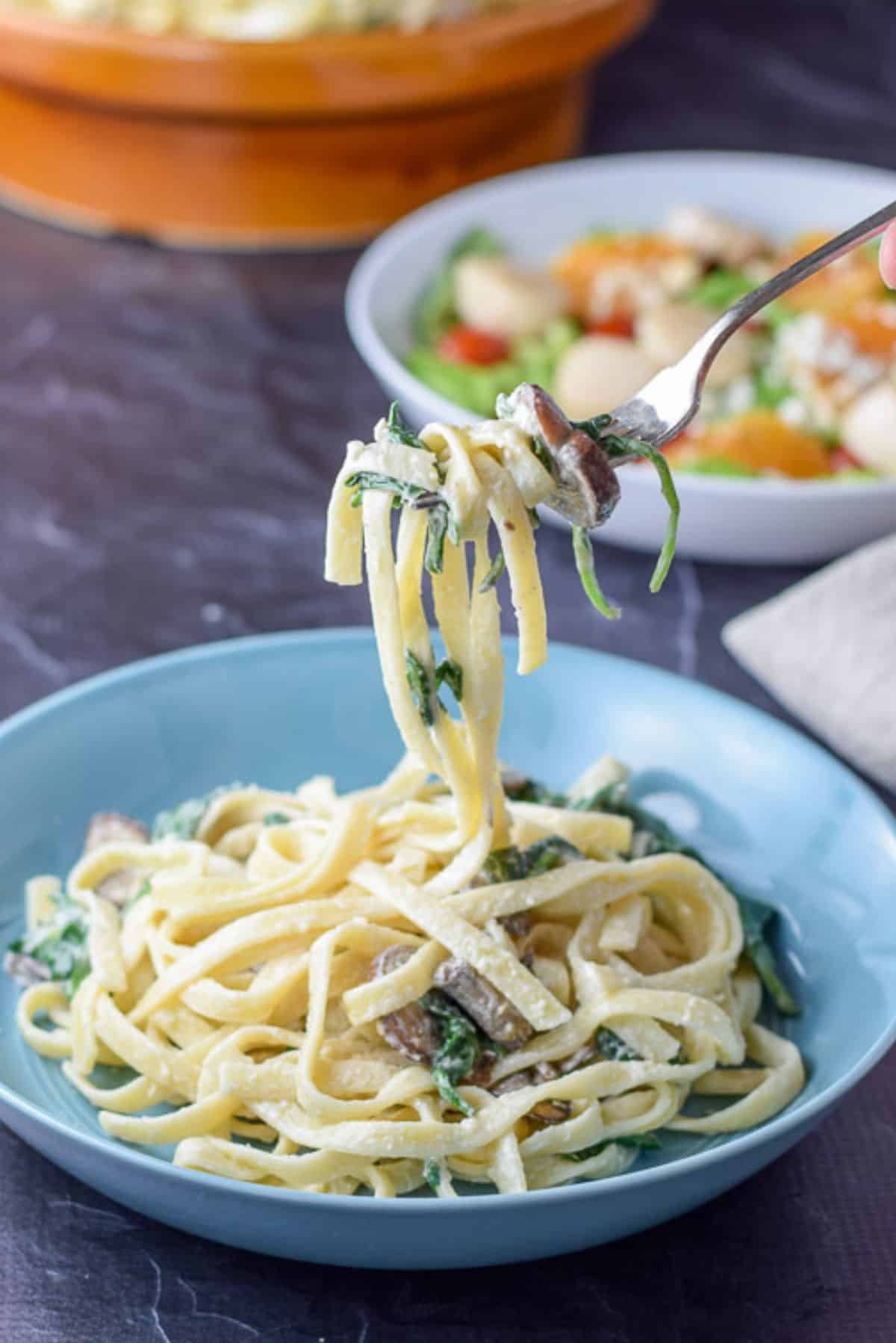A forkful of mushroom and fettuccine held above the shallow bowl with a salad and the big bowl in the background