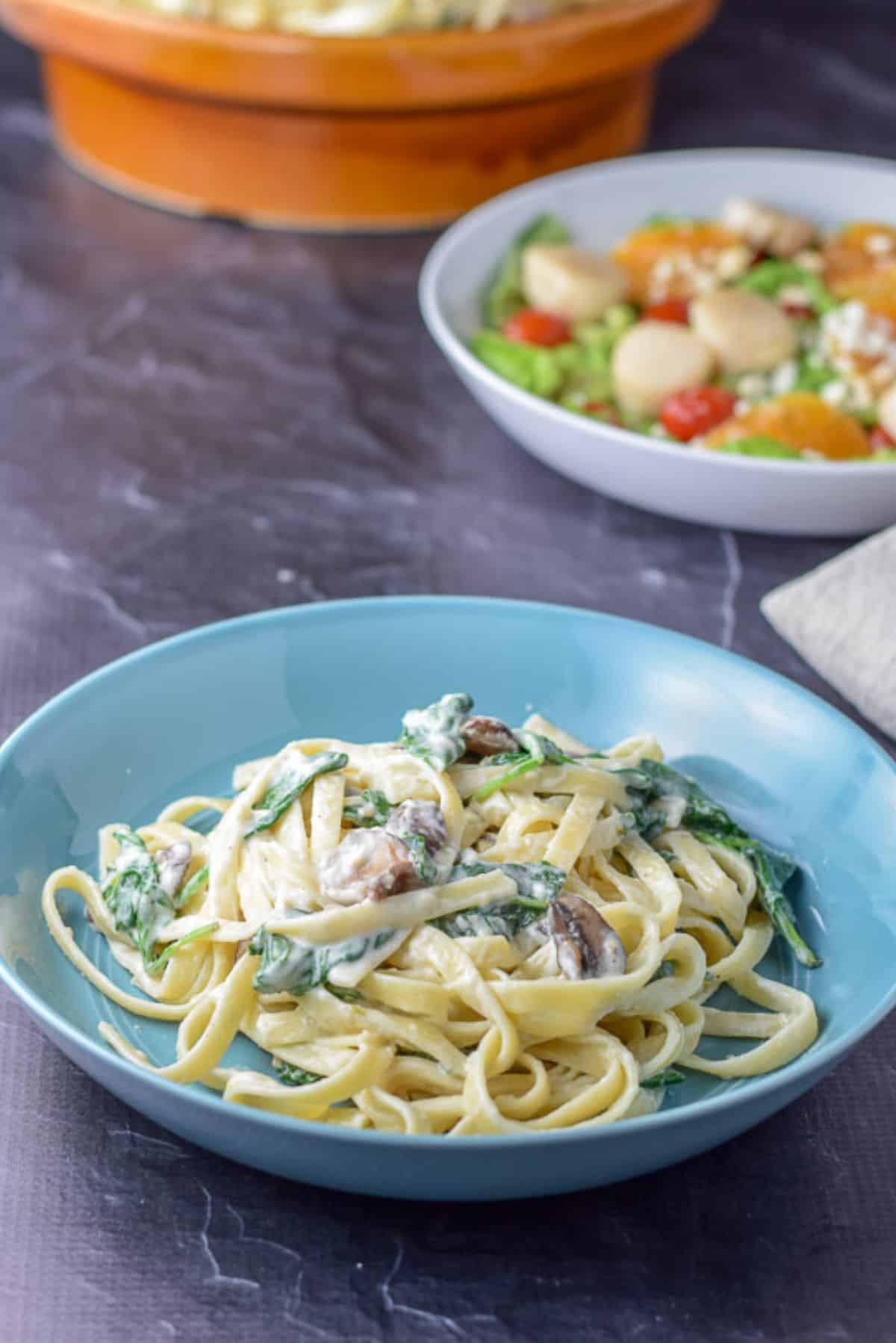 A blue shallow bowl with the fettuccine dish in it, there is a salad and the big bowl of fettuccine in the background