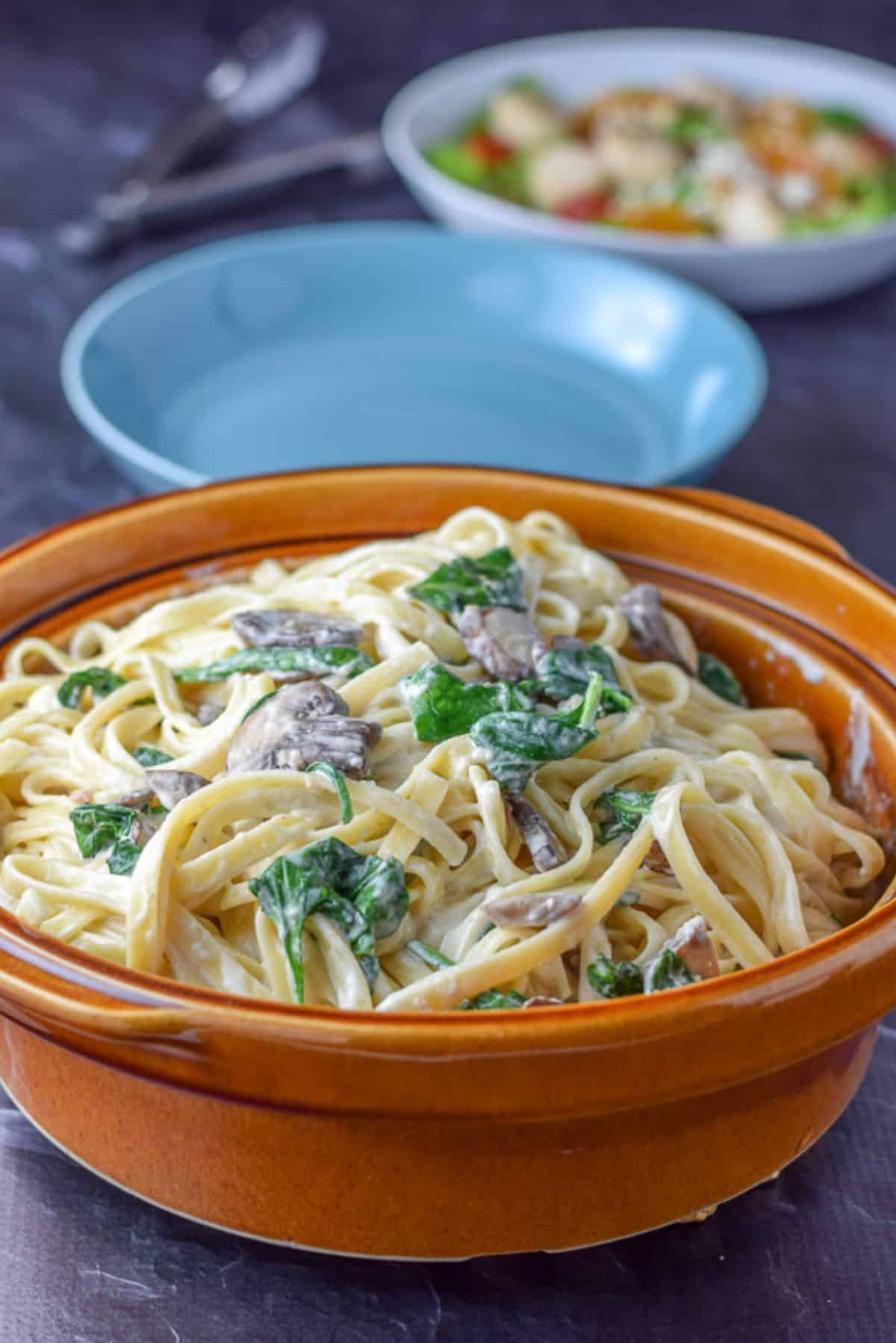 A big brown bowl of fettuccine with mushrooms and spinach with two bowls in the background