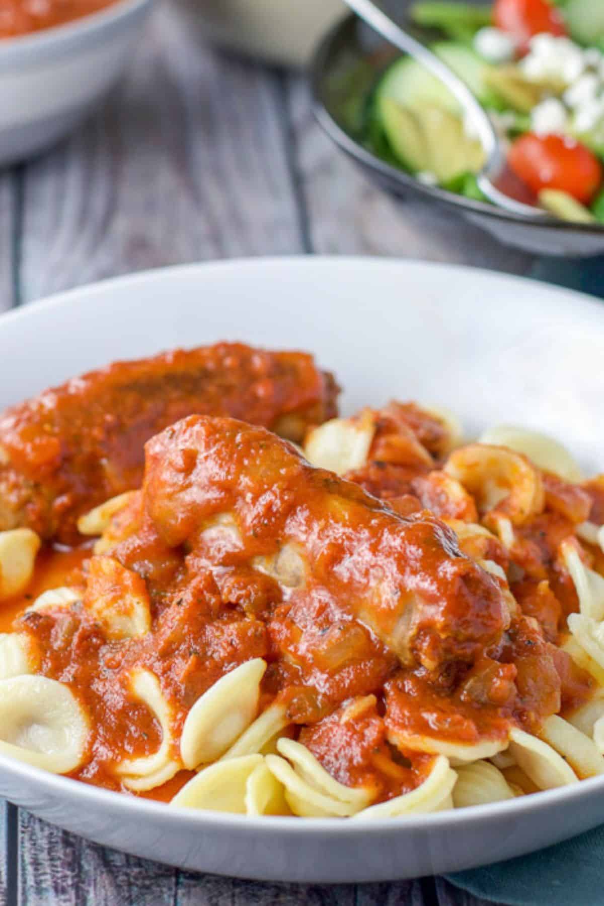 A sausage and gravy on some pasta in a white bowl. There is a salad and bowl of the sauce