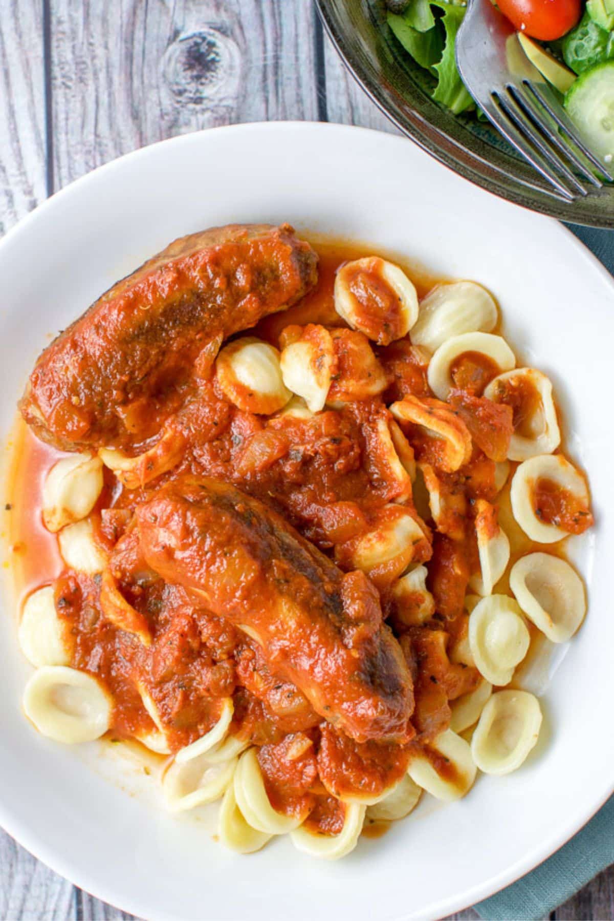 Overhead view of the a white bowl with two sausages with sauce and pasta
