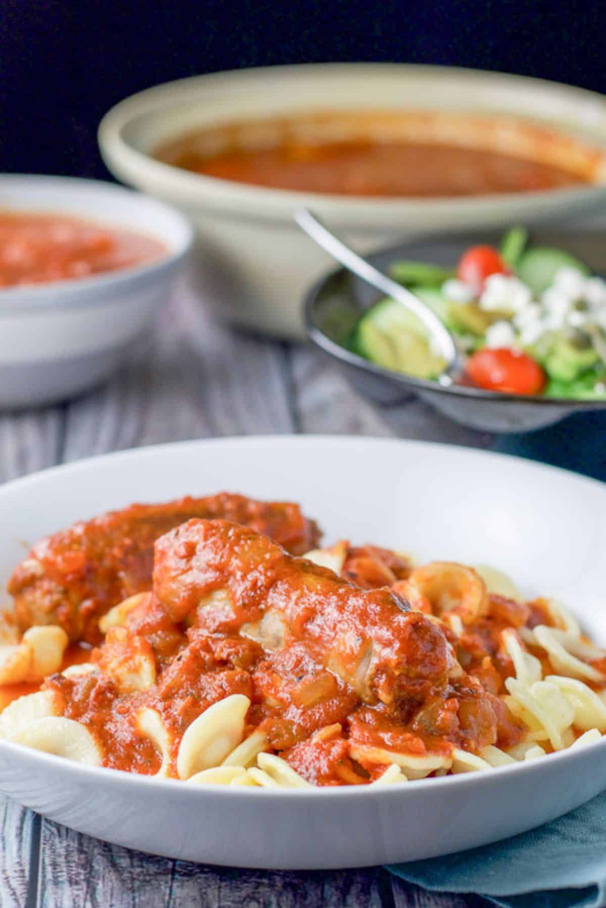 A white bowl filled with pasta with sauce and a sausage on top. There is a bowl with more sauce in the background