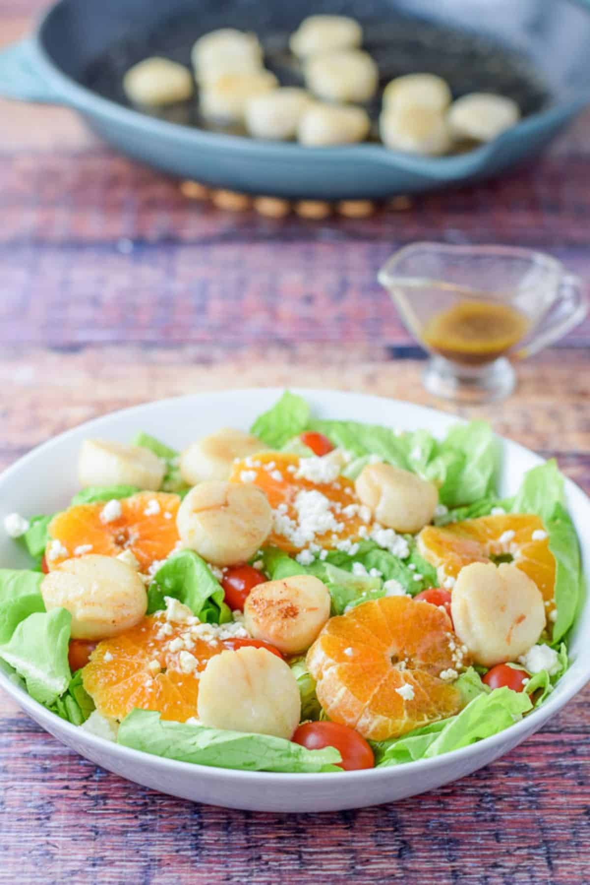 Dressing poured for on the scallop salad with the pan of scallops in the background with the dressing in the container
