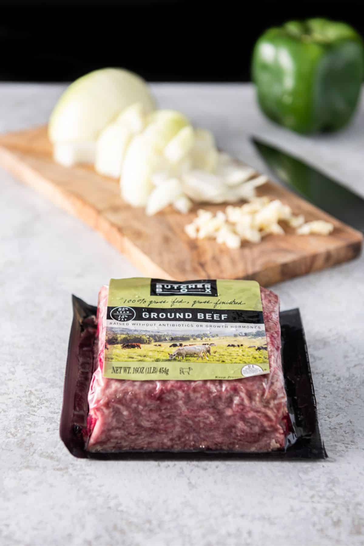 Ground beef in front with a board with a chopped onion and garlic on it with a bell pepper in the background