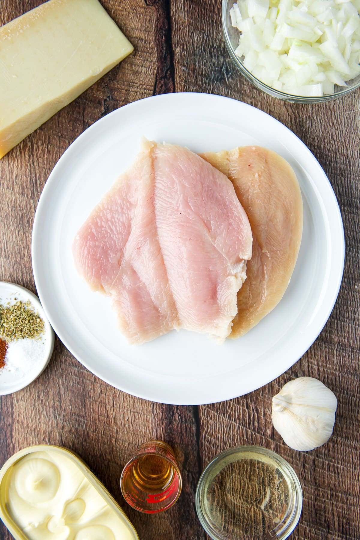 A white plate with filleted chicken breasts. Also an onion, cheese, herbs, and spices, butter, garlic, wine, and sherry.