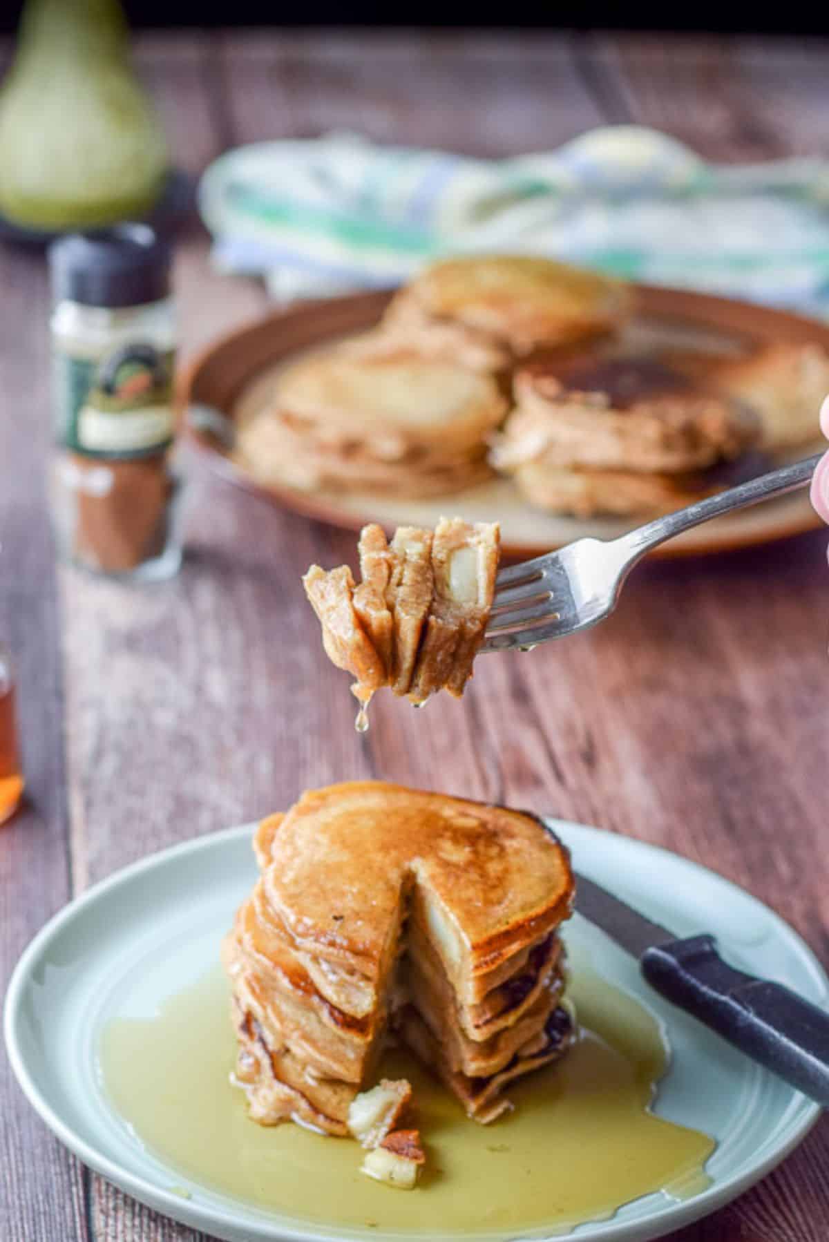 A hand holding a forkful of the pancakes. There is a plateful of pancakes in the background