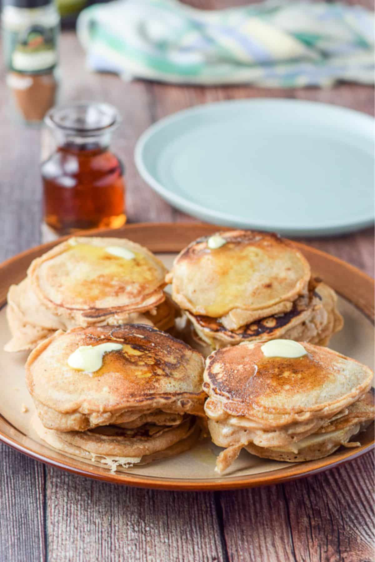 A brown plate with 4 stacks of pancakes and butter on them. There is a green plate, maple syrup and spices in the background