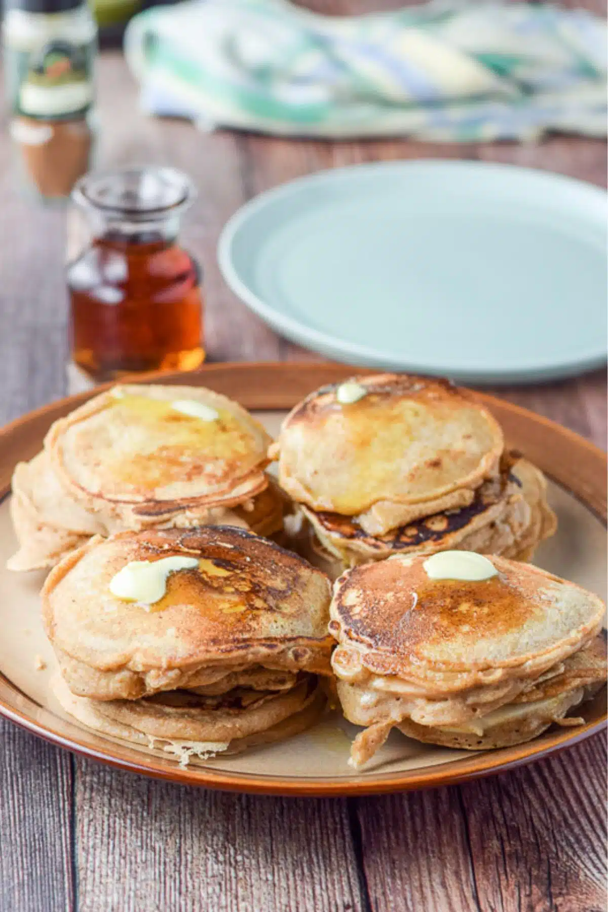 A brown plate with 4 stacks of pancakes and butter on them. There is a green plate, maple syrup and spices in the background