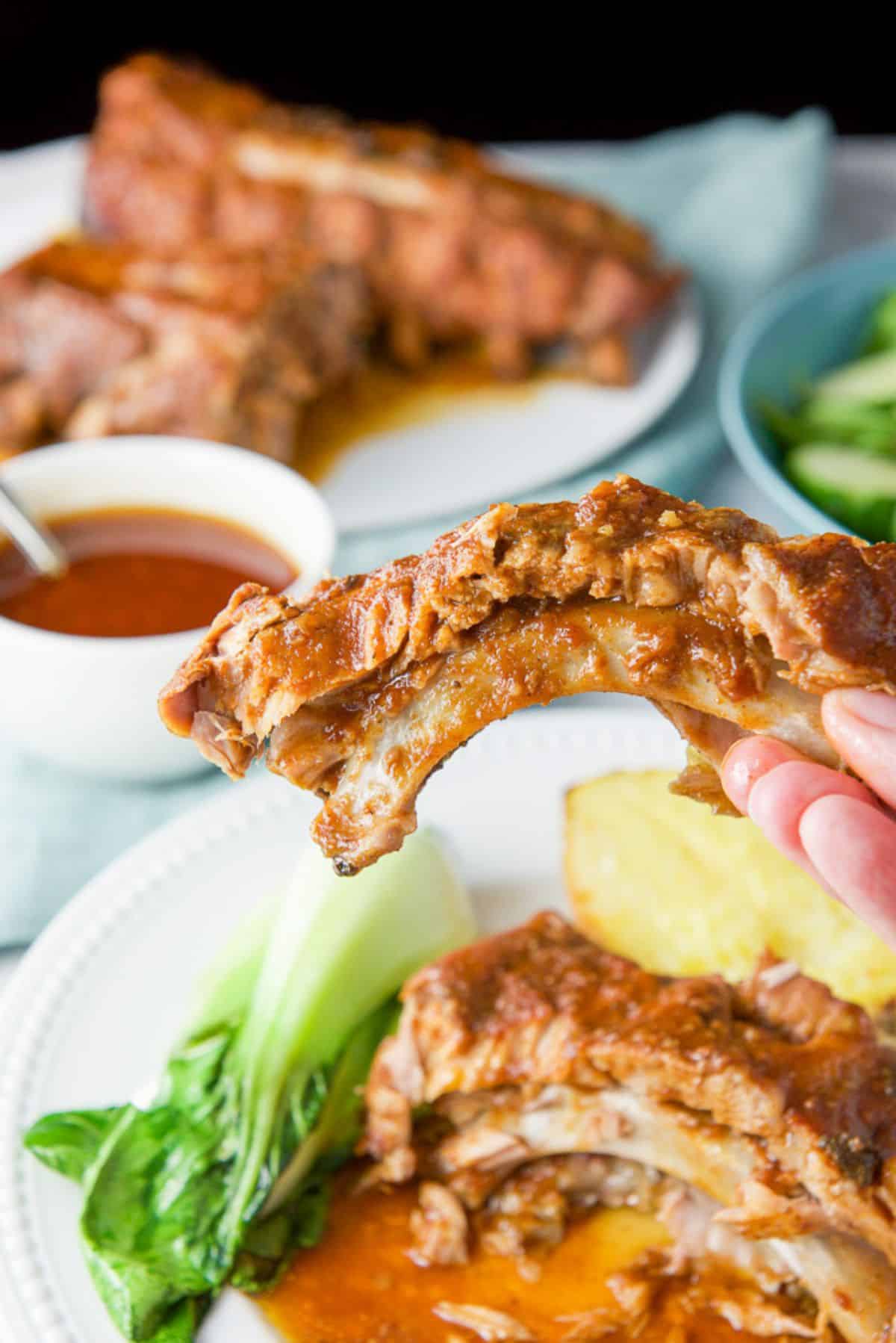 A hand holding one of the ribs over a plate of them. There are more ribs and sauce in the background