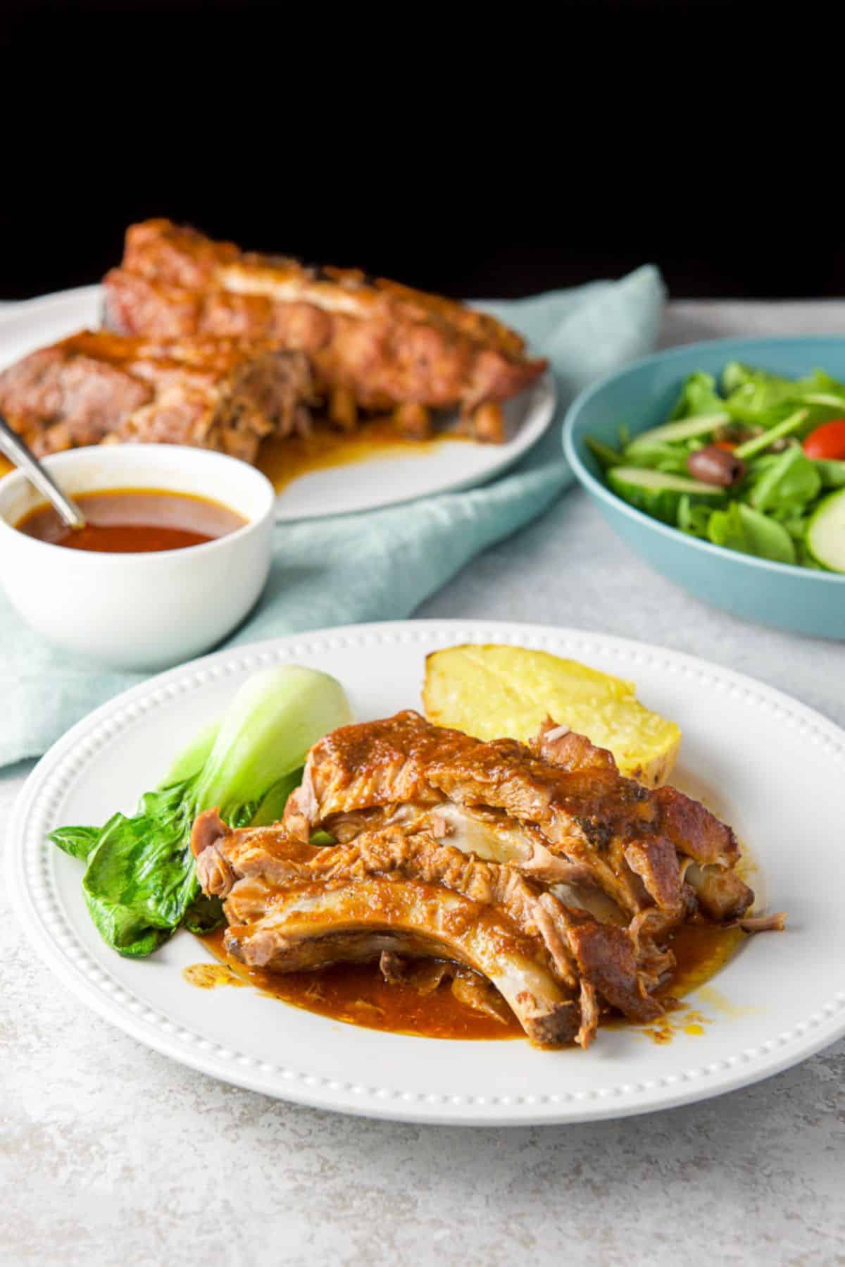 baby back ribs on a plate with potato and veggie. There is a platter of ribs, a salad and sauce in the background