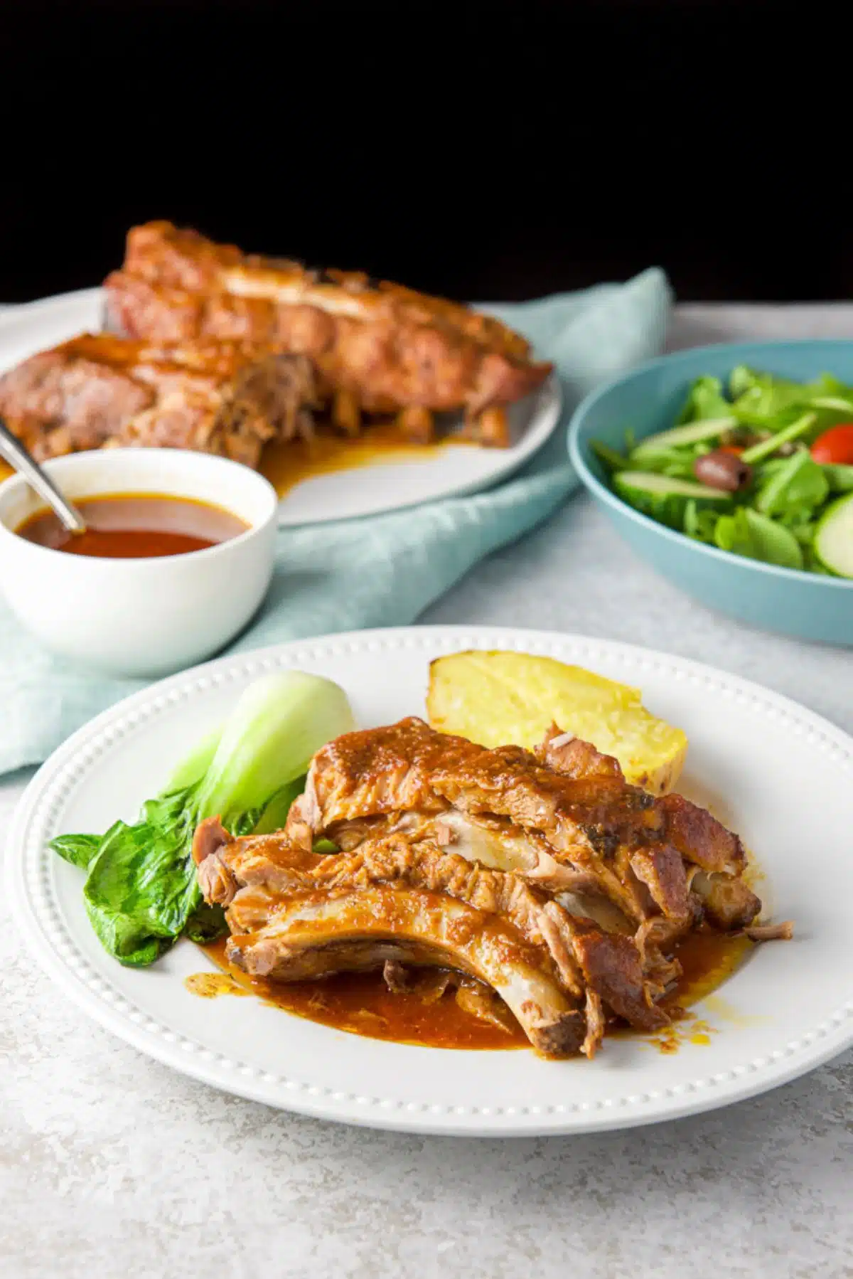 baby back ribs on a plate with potato and veggie. There is a platter of ribs, a salad and sauce in the background
