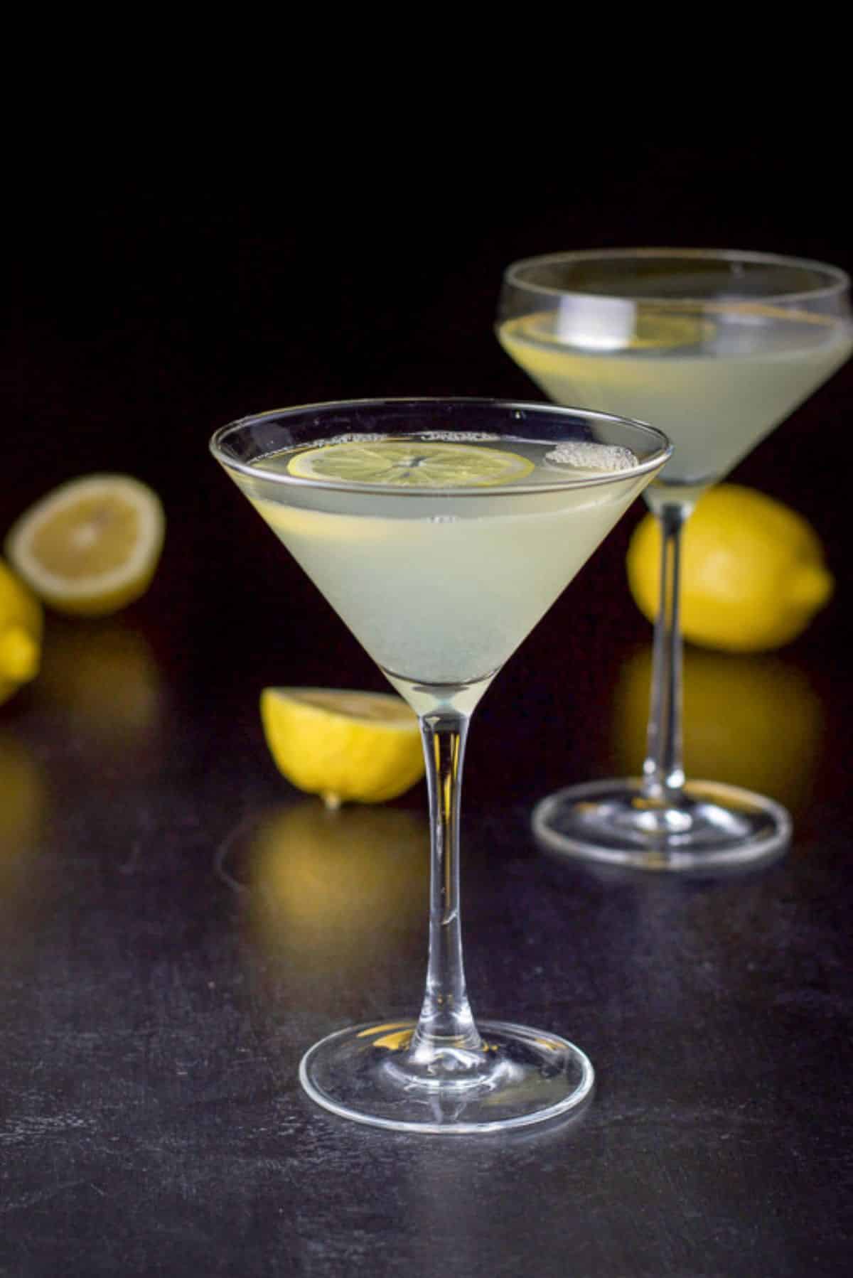 A classic martini glass filled with the lemon drink with the curved martini glass and lemons in the background