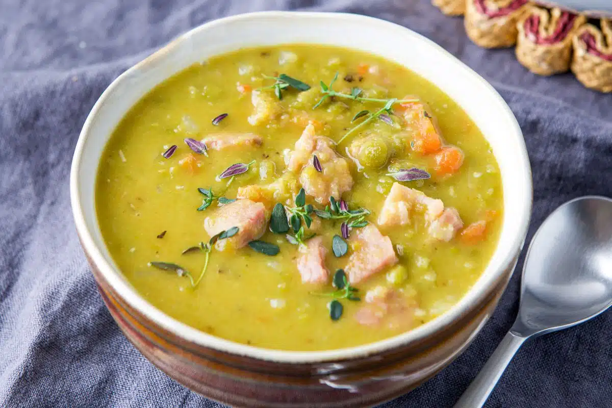 A brown bowl with a white inner part with pea soup in it, there is a spoon off the the right