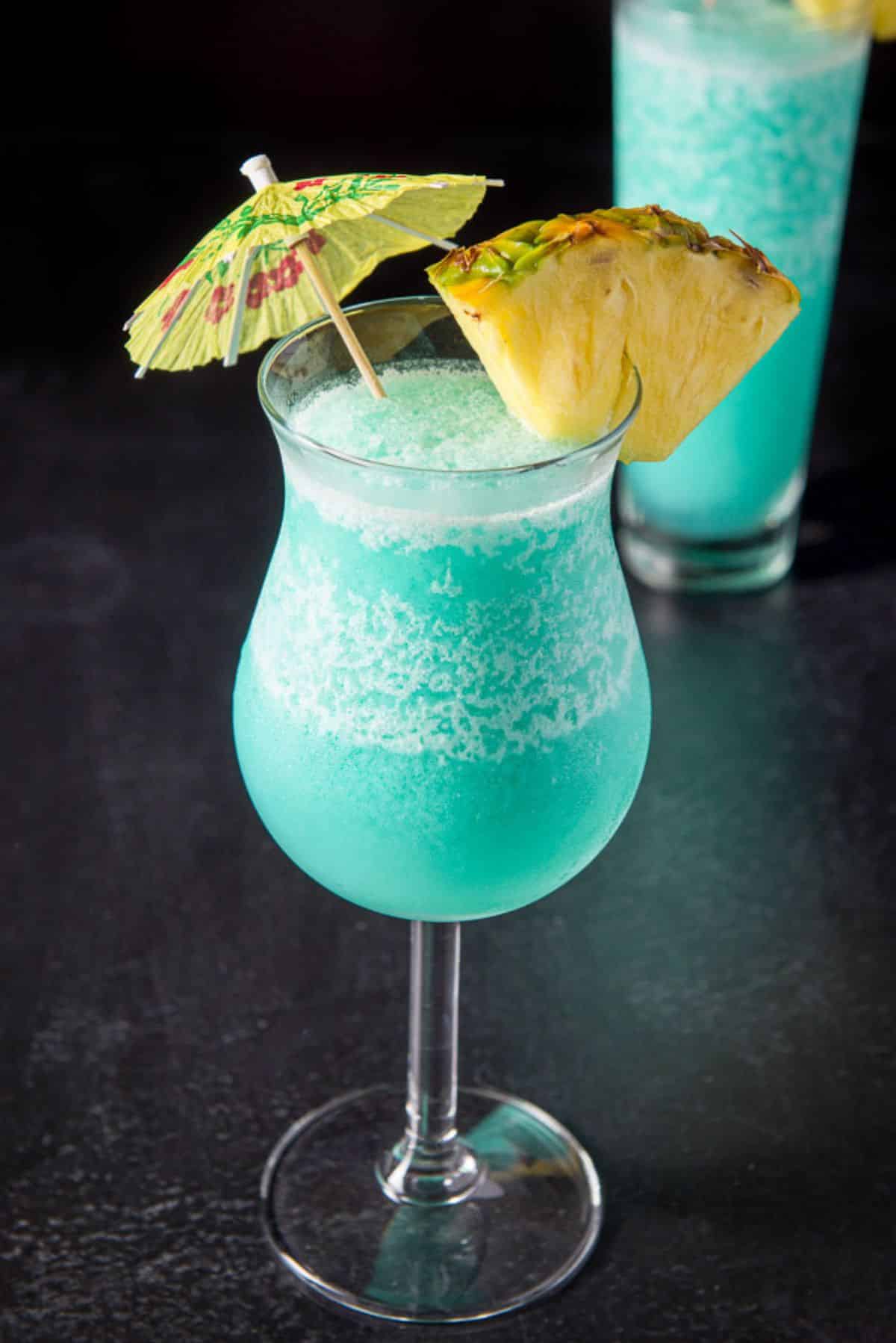 The tulip glass with the blue drink in front of the flared glass - garnished with pineapple and umbrellas