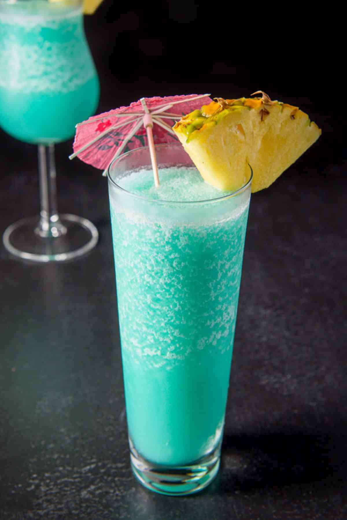 Flared glass filled with the frozen drink in front of the tulip glass. They are garnished with a pineapple wedge and umbrellas