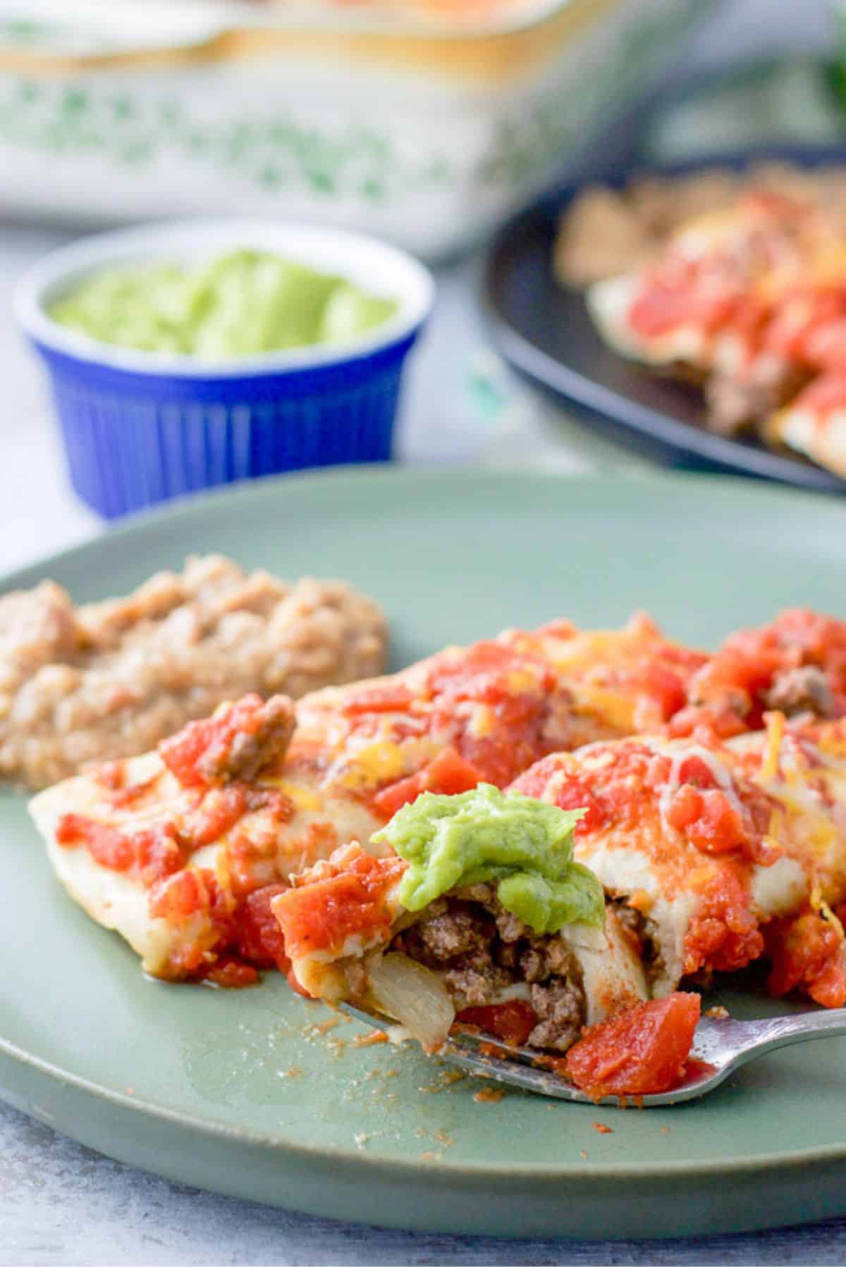 A fork with a bite of enchilada resting on a plate with two enchiladas and beans. There is guac and more enchiladas in the background