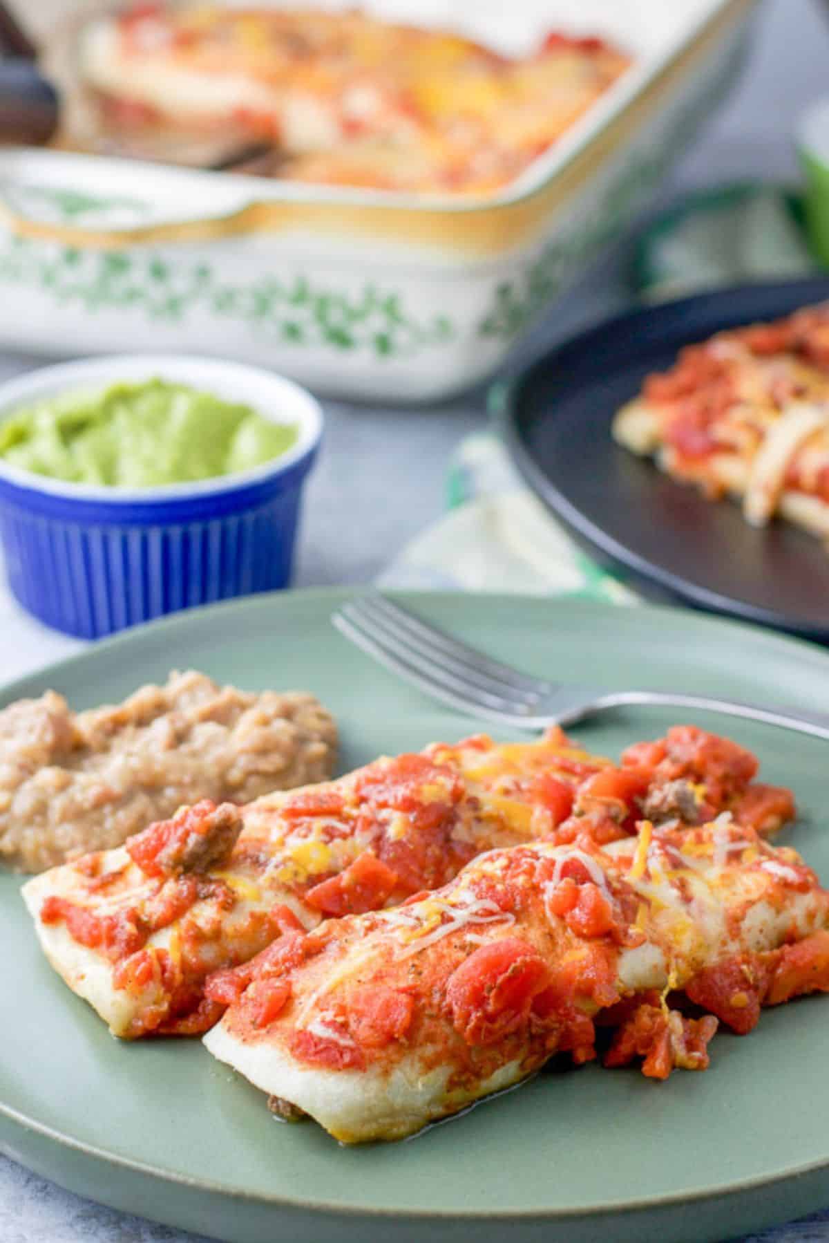 Two enchiladas with a red sauce on a plate with some beans. There is some guacamole in the background along with another plate of enchiladas