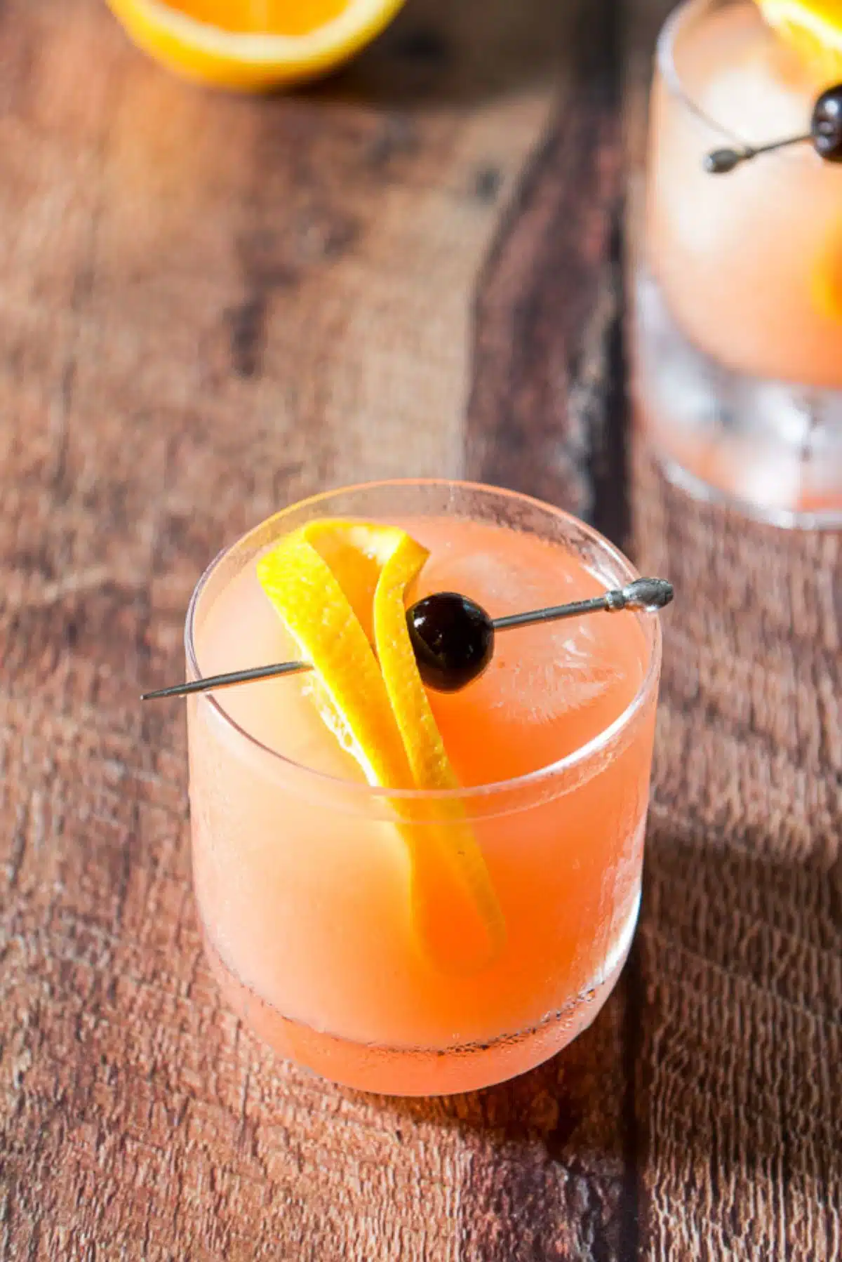 Close view of an apricot colored cocktail with a skewer with a bended orange slice and cherry