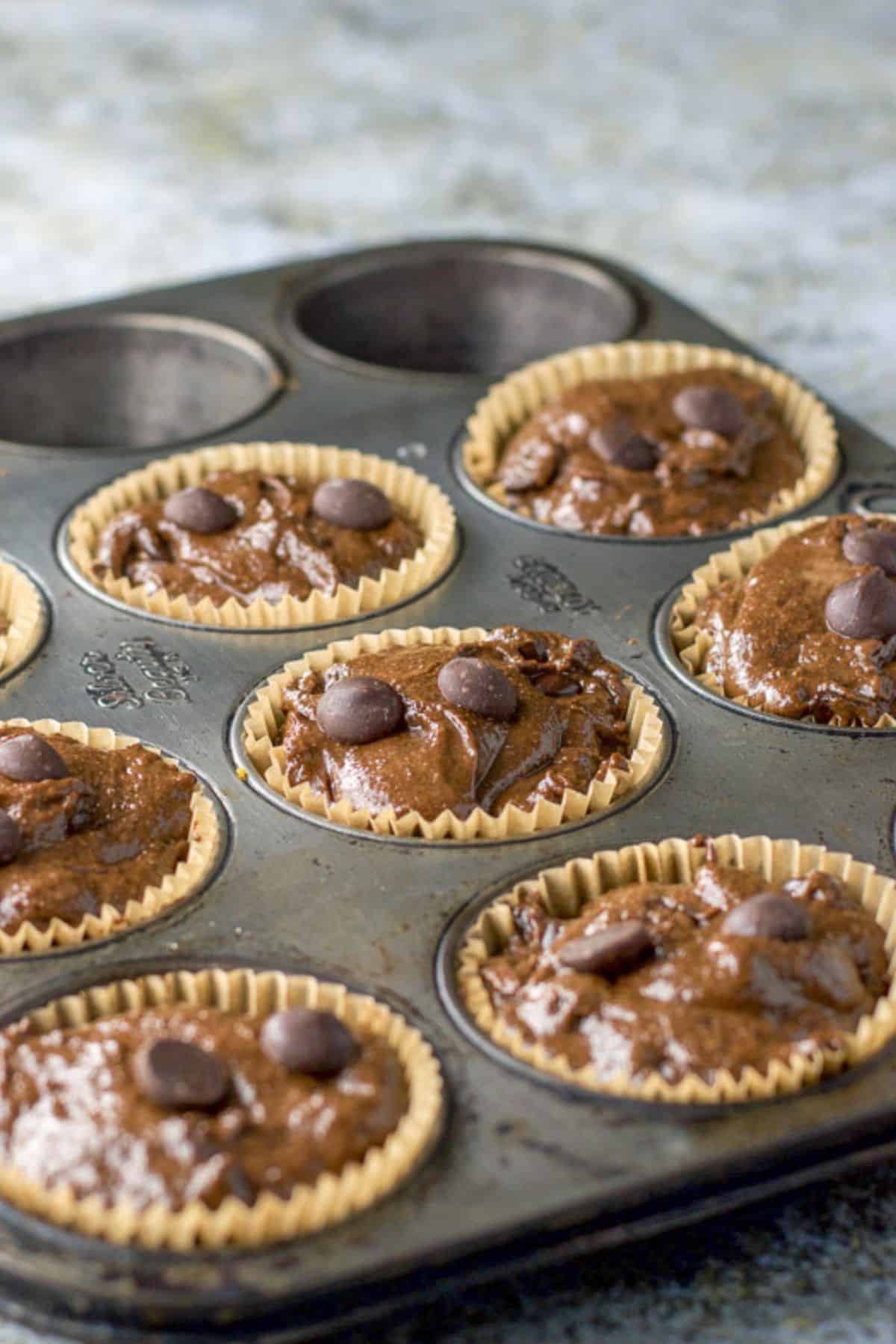 A muffin tin with nine baking cups in it along with the chocolate muffin batter and chocolate chips