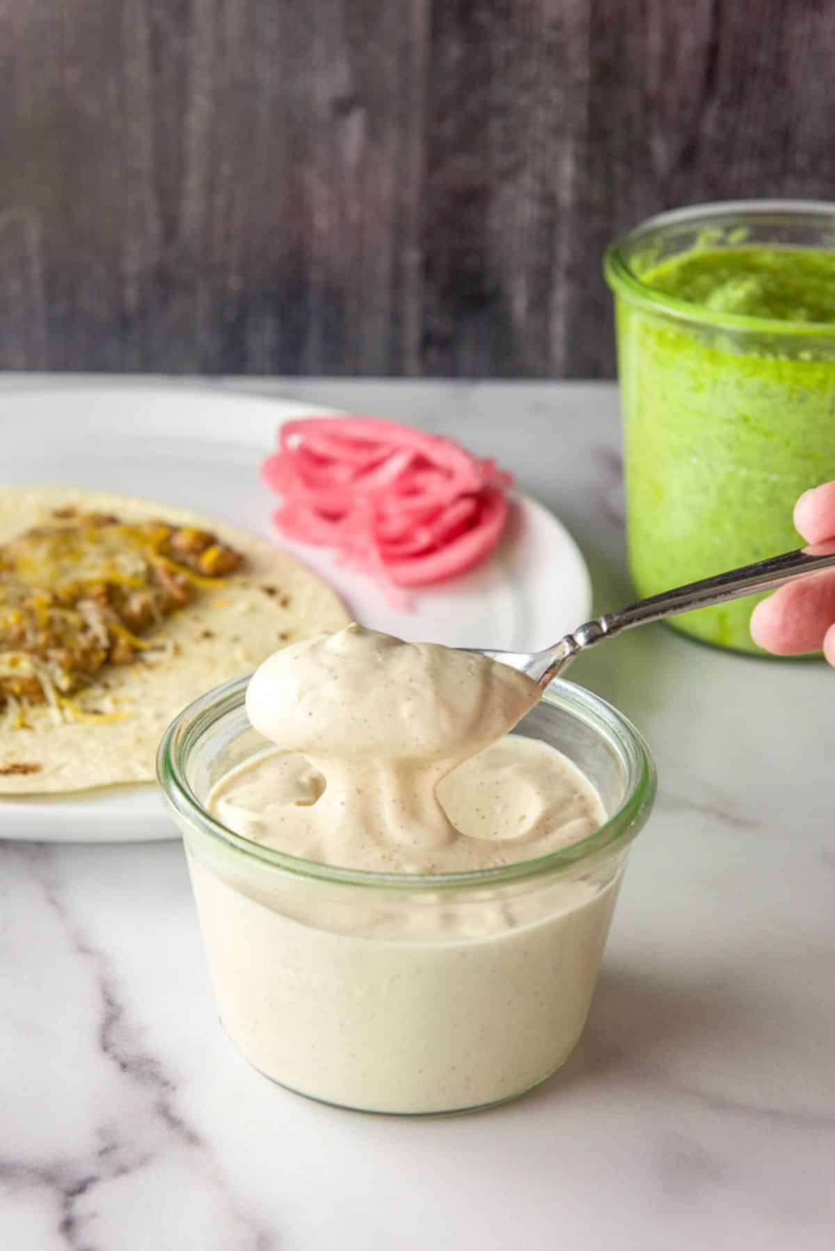 Spoonful of Mexican crema over the jar with a green sauce and plate of food behind