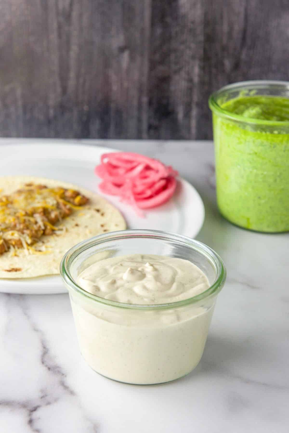 Crema in a glass jar ready to be used in front of a plate with pickled onions, tortilla and green sauce