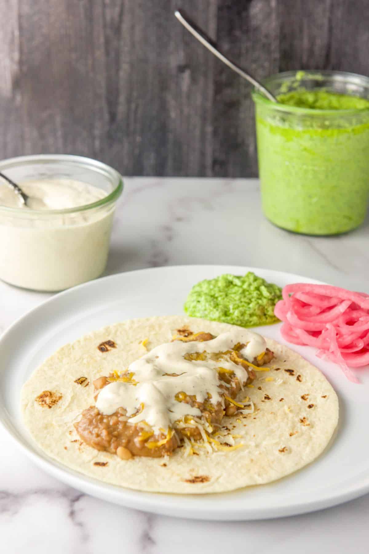 White sauce on some beans and cheese in a flour tortilla, jars of crema and green sauce in the back