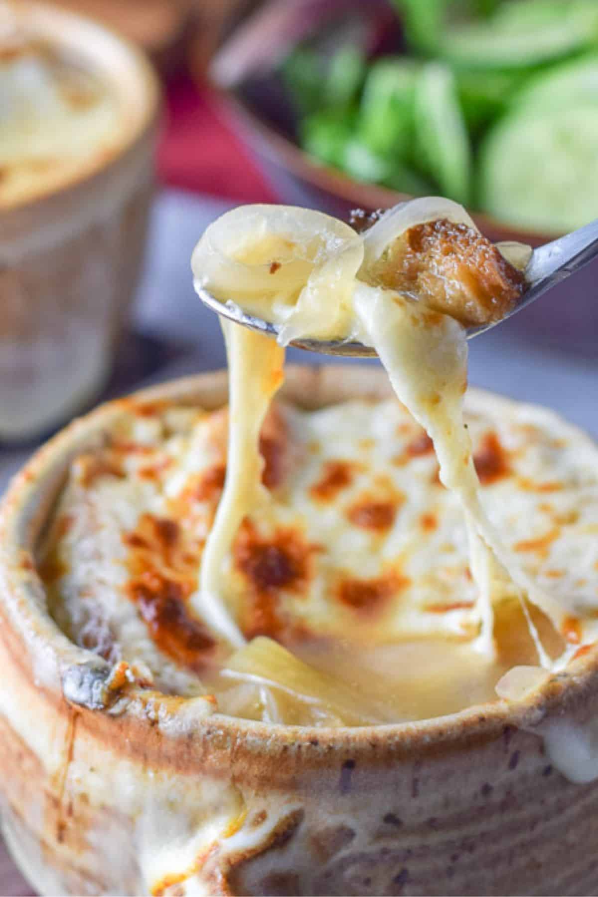 A cheesy spoonful of onion soup held over the crock with salad and bread in the background