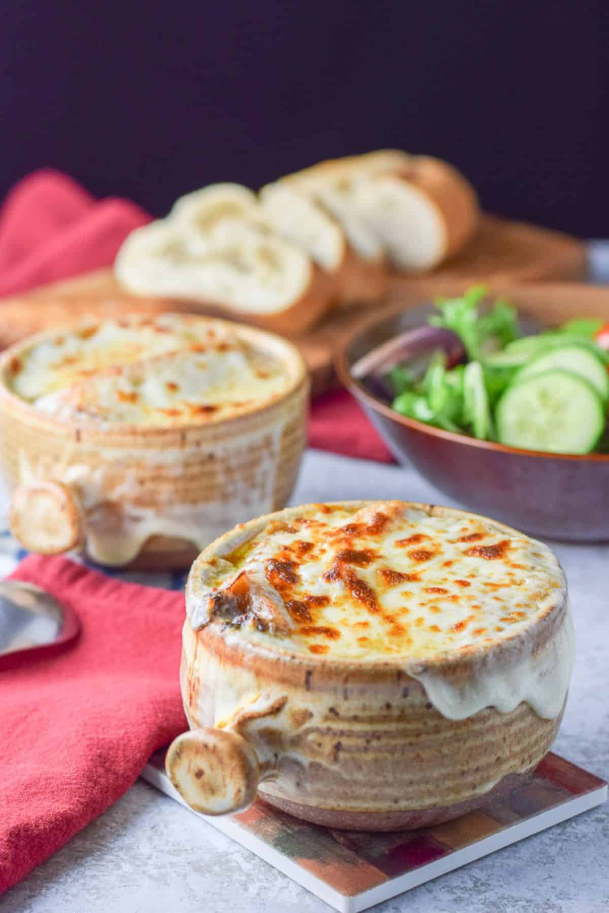 Cheese melted on the crocks of soup with a salad in the background and bread on a cutting board