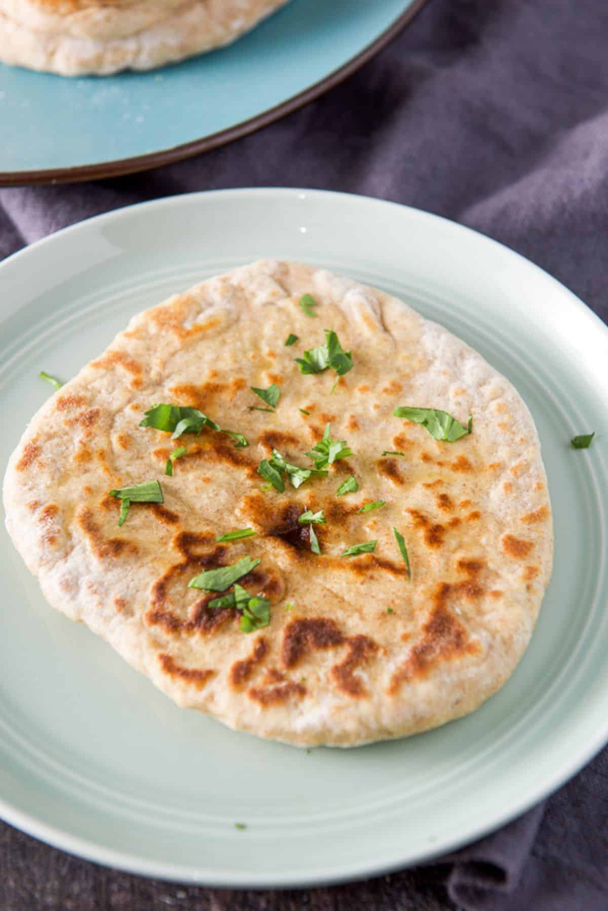 A naan round on a green plate with butter on it and cilantro. There is a pile of bread in the background