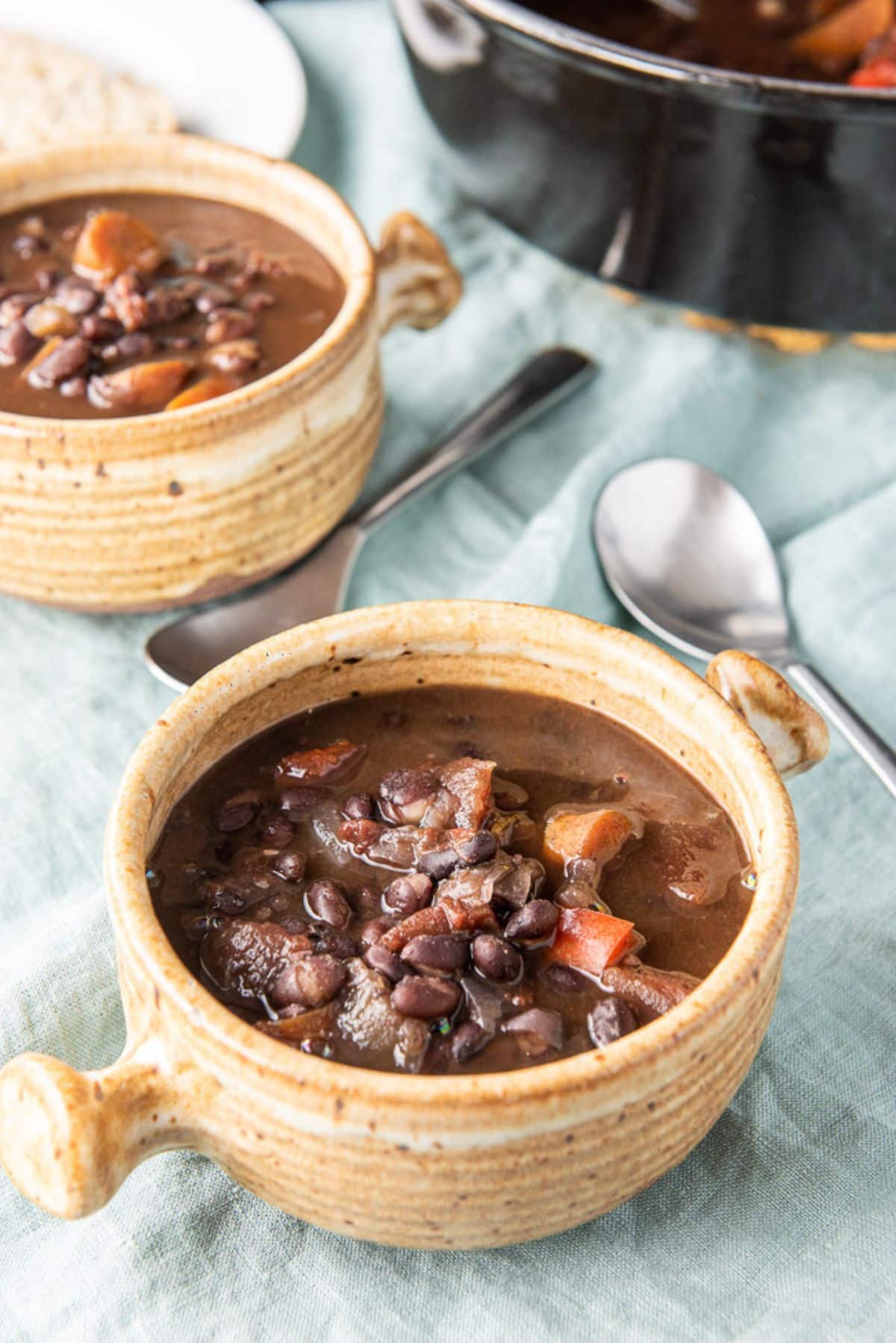 Close up of two brown crocks of bean soup with the spoons and pan in the background