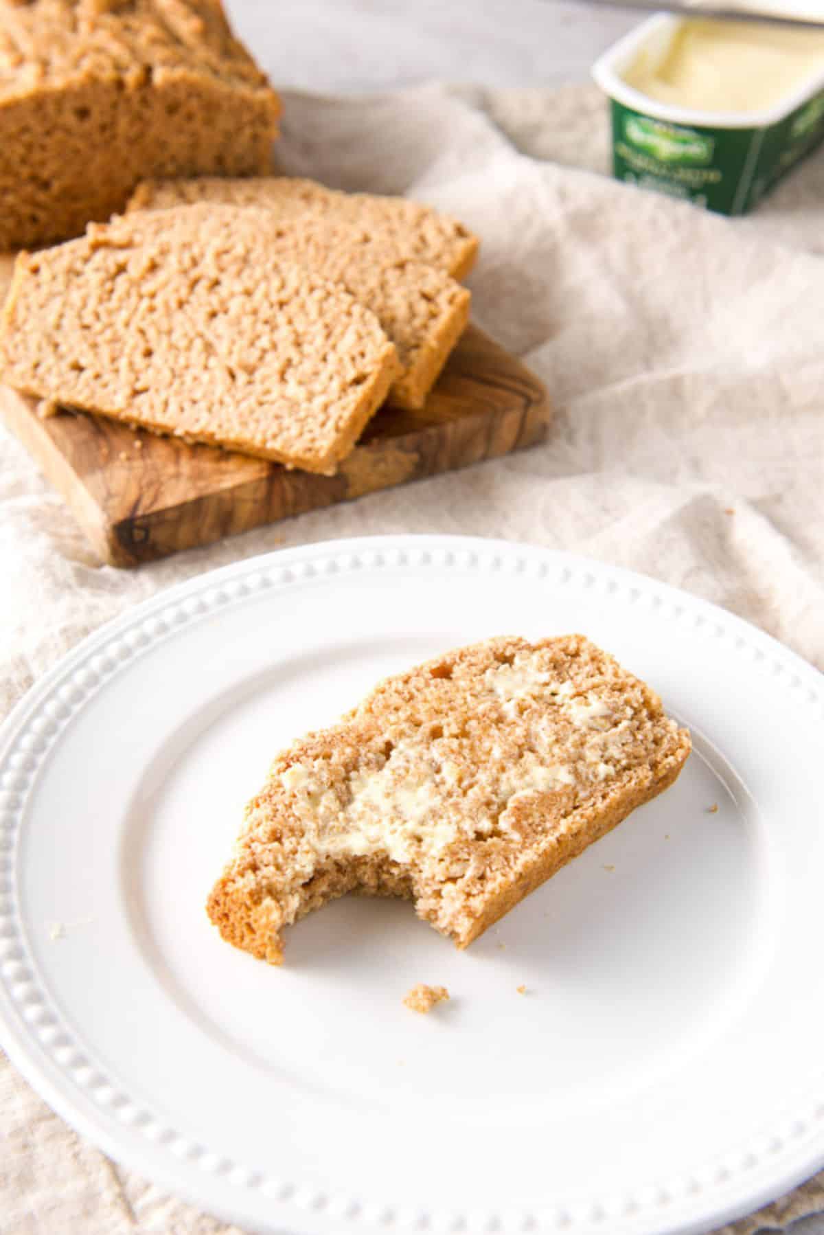 A white beaded plate with a slice of beer bread on it, with butter and a bite taken out of it. More slices and butter in the back