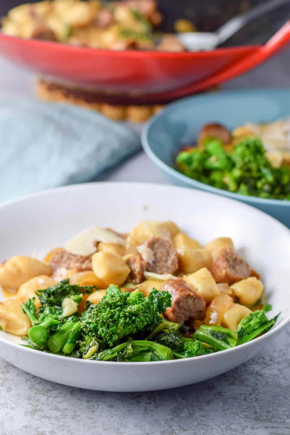 A deep white dish with the rabe and gnocchi in it with another dish and the wok in the back