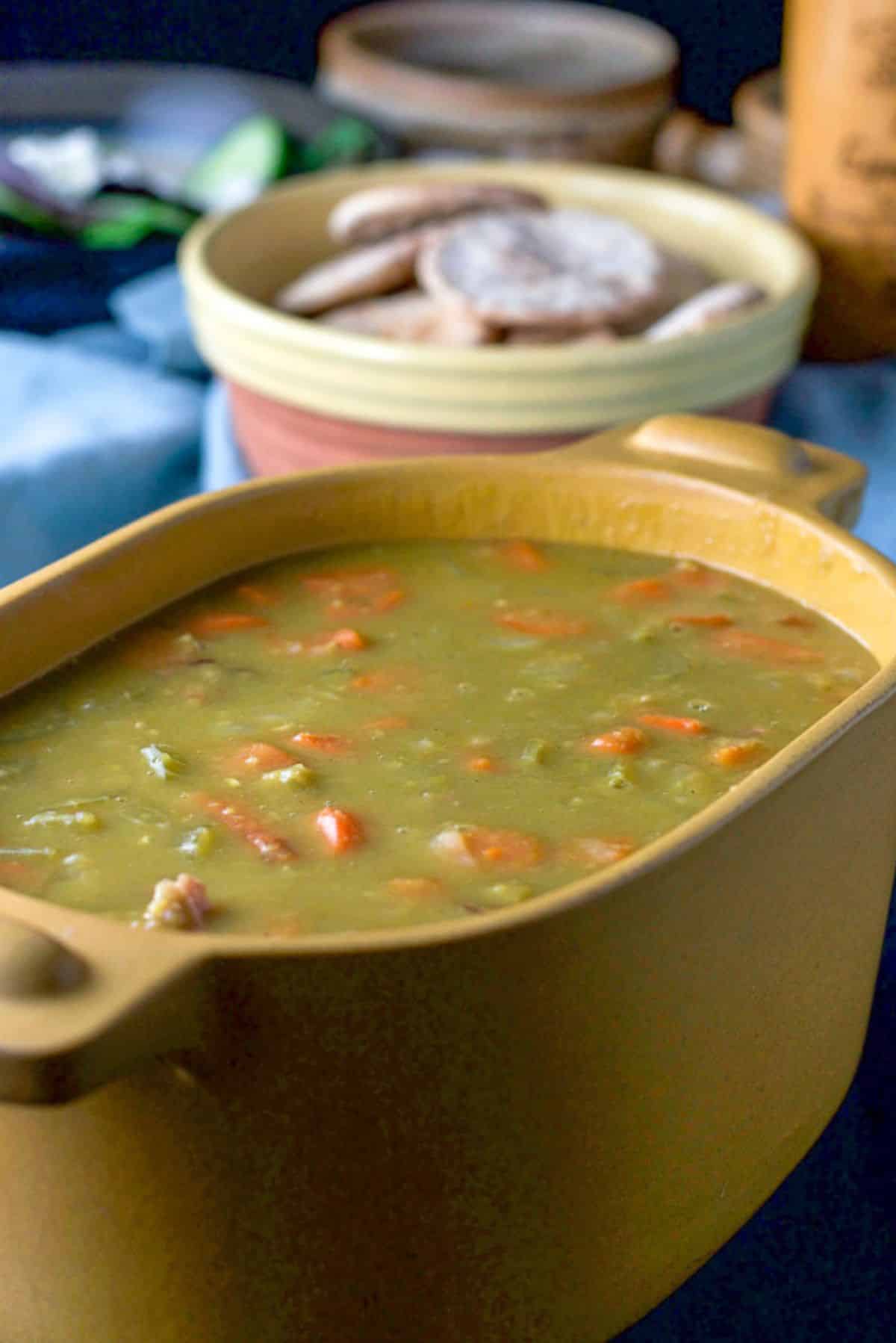A mustard colored casserole dish filled with pea soup. There are some pita bread in a bowl, a salad and a pepper mill in the background