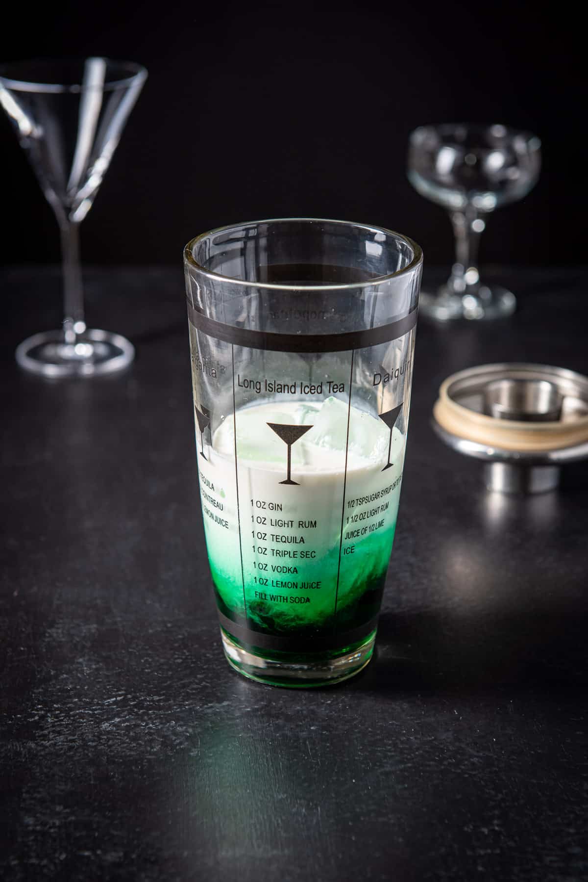 A shaker filled with a green liqueur, and cream with two glasses in the back