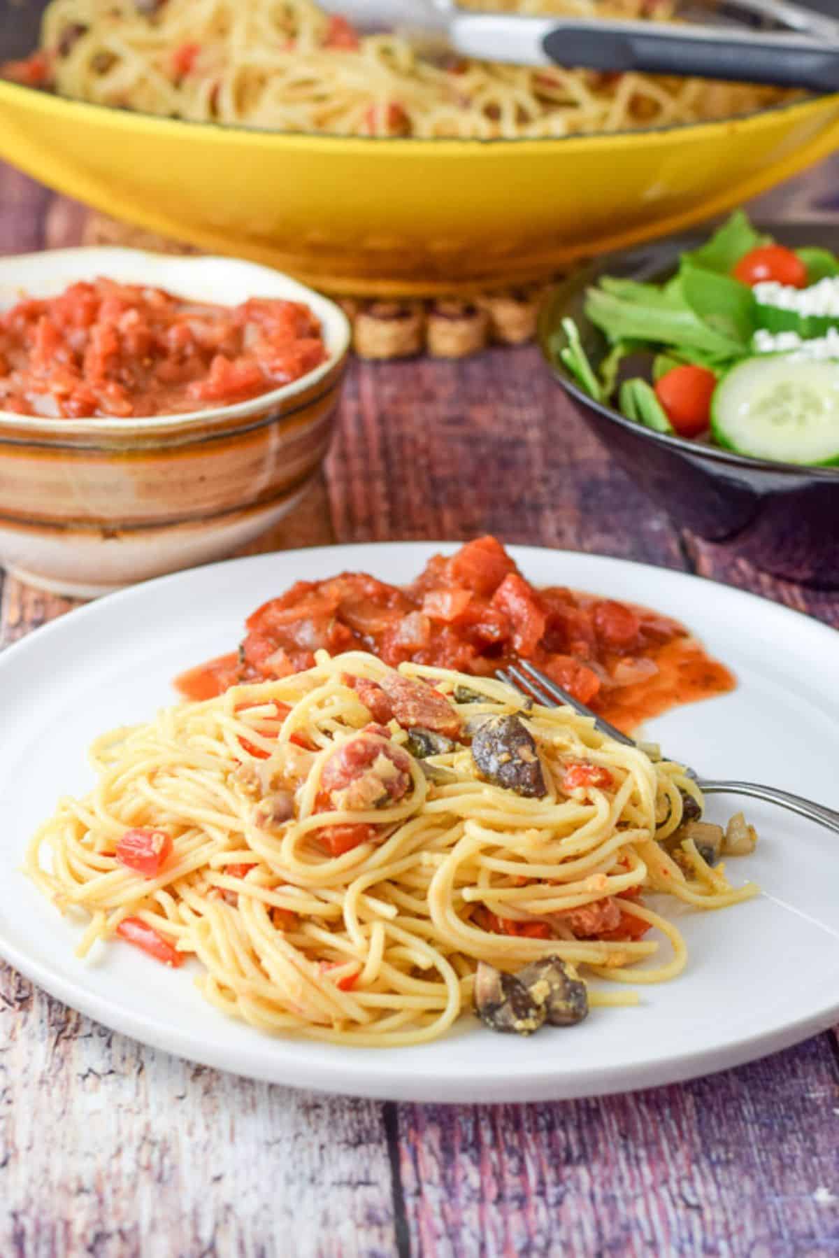 A fork on a plate with the pasta, sauce, and vegetables. There is a bowl of sauce and a wok filled with the meal in the back