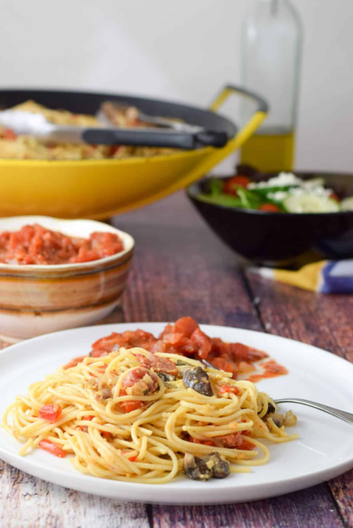 A white plate with the spaghetti on it with some sauce, salad, and wok filled with the spaghetti in the back