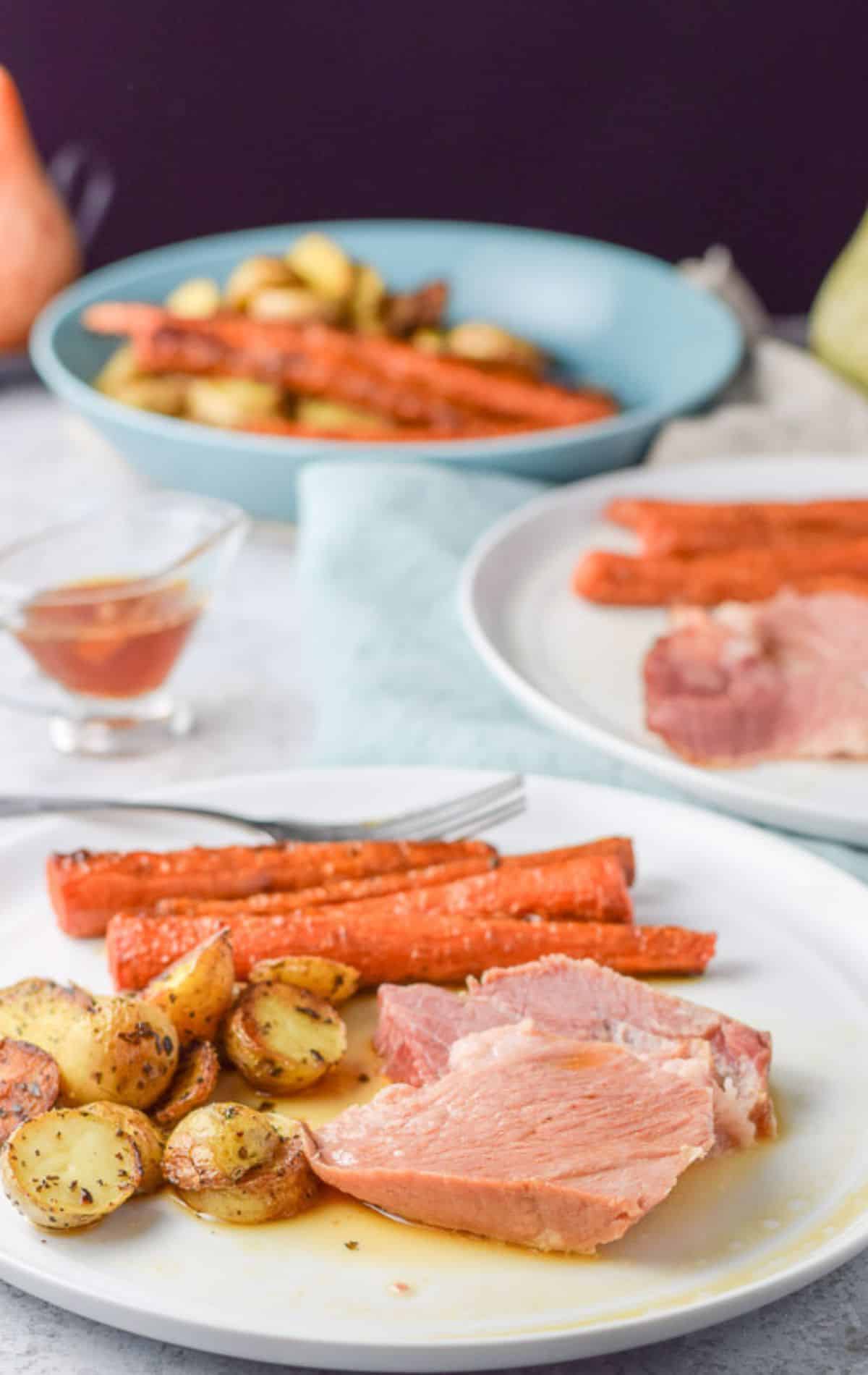 A white plate with meat, potato, and carrots in front of another plate and a bowl of vegetables